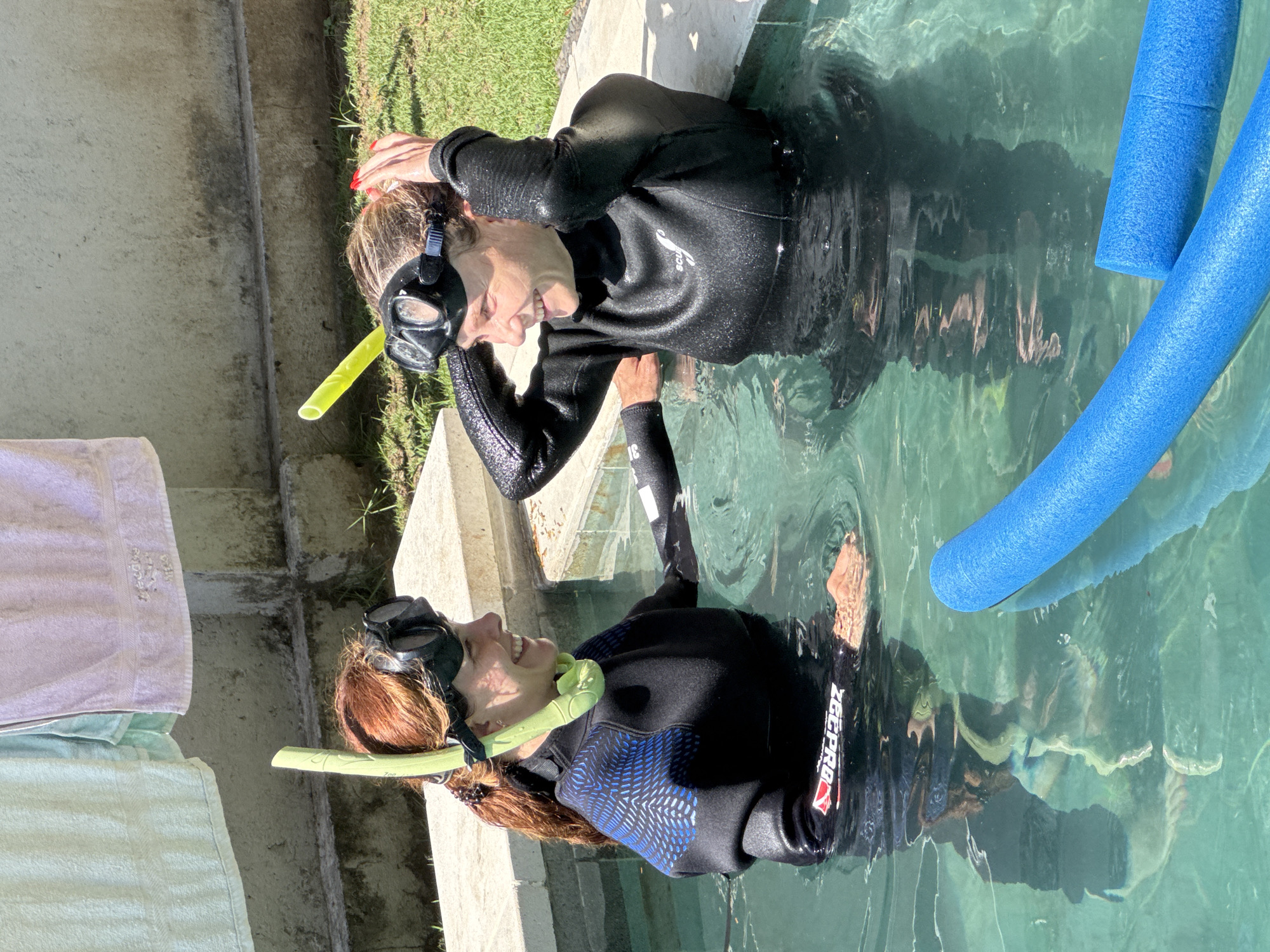 Two women in wetsuits and snorkels are in a pool of clear, turquoise water. The woman on the left has auburn hair and is smiling. The woman on the right has blonde hair and is adjusting her snorkel mask. Two blue pool noodles are visible in the water. In the background, there are concrete structures, green grass, and laundry hanging on a clothesline. The scene is set in Nusa Penida, Indonesia, likely in a private residence with a pool.
