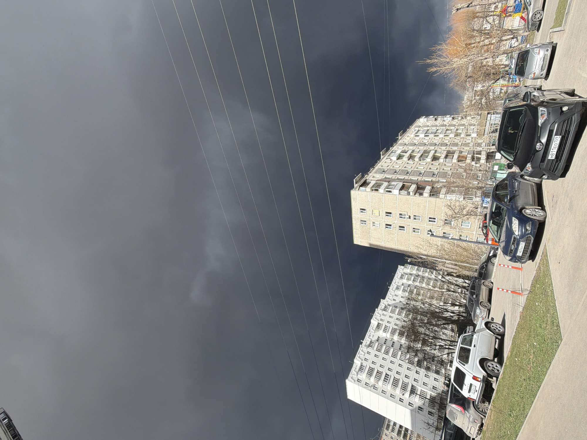 The image depicts an urban street scene in Perm, Russia, under a heavily overcast and dark sky. The sky, dominating the upper portion of the frame, is a dramatic dark grey to almost black, suggesting either an impending storm or recent adverse weather. Multiple thin power or communication lines stretch diagonally across the sky from the upper left to the lower right.
In the lower half of the image, several multi-story residential buildings, characteristic of Soviet-era architecture with numerous windows and balconies, line a street or parking area. Their light-colored facades contrast sharply with the dark sky. A row of parked cars occupies the foreground and mid-ground. Visible vehicles include a black SUV displaying license plate M811EO 159, a blue sedan with plate T483KH 59, and a silver hatchback with plate E021HH 59. The "59" region code on these plates confirms the location as Perm Krai, Russia. A white SUV with plate A777TT is also visible.
Between the buildings and cars, there are patches of green grass and several bare-branched trees, indicating either early spring or late autumn. A small, colorful playground structure is discernible in the background among the trees. The scene is static, with no people visible or specific activities taking place. The overall mood is quiet and somewhat somber due to the heavy cloud cover.