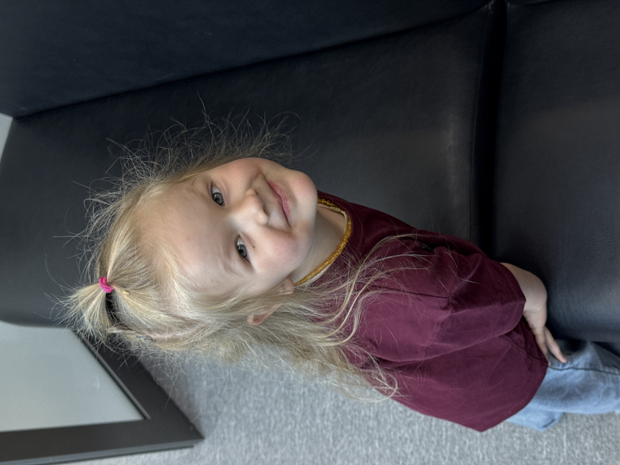 A young girl with blonde hair pulled back into a small ponytail with a pink hair tie is lying on a black leather couch. She is wearing a maroon long-sleeved shirt and blue jeans. She is smiling and looking directly at the camera. She is wearing a yellow beaded necklace. The background consists of a gray carpet and a black framed object. The photograph was taken in Perm, Russia.