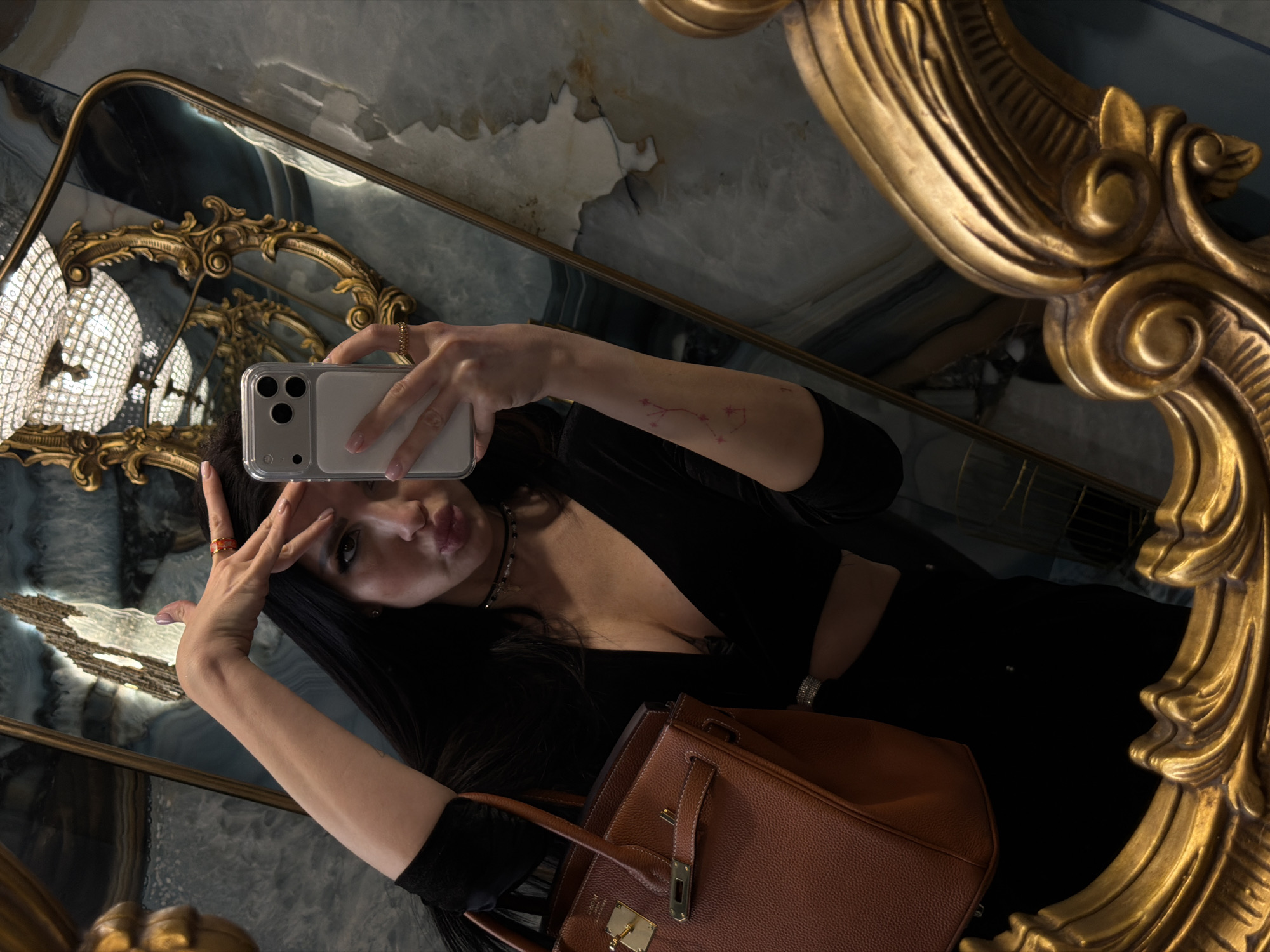 A female subject is captured in a mirror selfie within an opulent restroom at Café Pushkin in Moscow, Russia. The subject, with long dark hair, wears a black, long-sleeved, low-cut top revealing her midriff, a thin black choker, and a diamond bracelet on her left wrist. She has light pink fingernails, a golden ring on her left ring finger, and another on her right ring finger, which is raised to her forehead. A small, red, constellation-like tattoo is visible on her right forearm. She holds a silver-colored smartphone with a clear case in her left hand, pointing it towards the mirror to take the photo, while her lips are pursed. A brown, textured leather handbag with prominent gold hardware (consistent with a Hermes Birkin) hangs from her left arm. The environment features highly polished walls composed of light blue and white veined marble or onyx. A large, ornate gold-framed mirror occupies the foreground, reflecting the subject, while a similar gold-framed mirror in the background reflects a large, multi-tiered crystal chandelier.