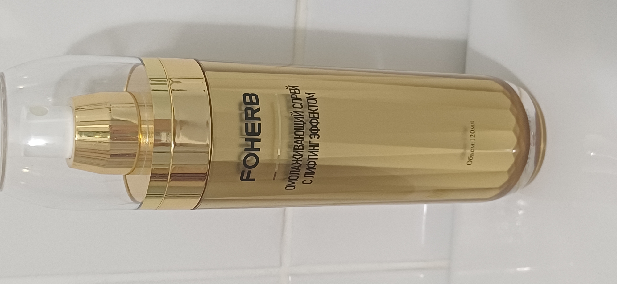 A close-up, eye-level, full-shot image shows a gold-colored spray bottle on a white tiled surface. The bottle has a clear plastic cap that covers a gold pump mechanism. The brand name "FO HERB" is printed in black text on the front of the bottle, below which is text in Russian that reads "ОМОЛАЖИВАЮЩИЙ СПРЕЙ С ЛИФТИНГ ЭФФЕКТОМ" (Rejuvenating spray with lifting effect). At the bottom of the bottle, also in black text, is "Объем: 120мл" (Volume: 120ml). The background is a white tiled wall. The image does not depict any people or indicate a location in Ekibastuz, Kazakhstan.