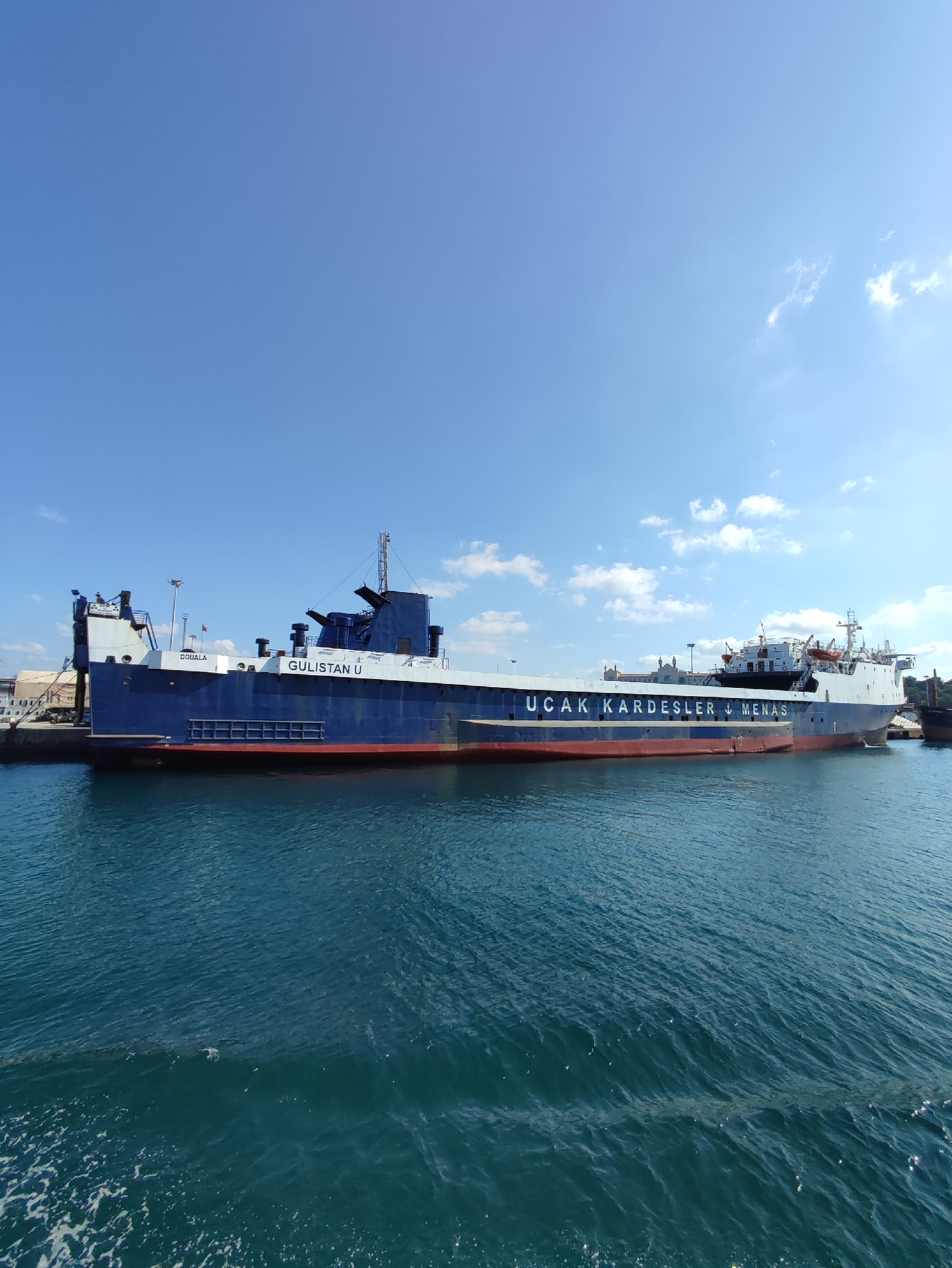 The image shows a ferry boat docked at a harbor. The boat is large and blue with red trim along the bottom. The name "GULISTAN U" is visible on the side of the boat, along with other lettering that is partially obscured. The boat is in the water, and the water is a deep blue color. The sky is clear and blue with a few scattered clouds. The scene appears to be in a city, with buildings visible in the background. The overall impression is of a sunny day in a busy port. The provided crops indicate that the location is Istanbul, Turkey, and that this ferry is named "UÇAK KARDEŞLER - MENAS".