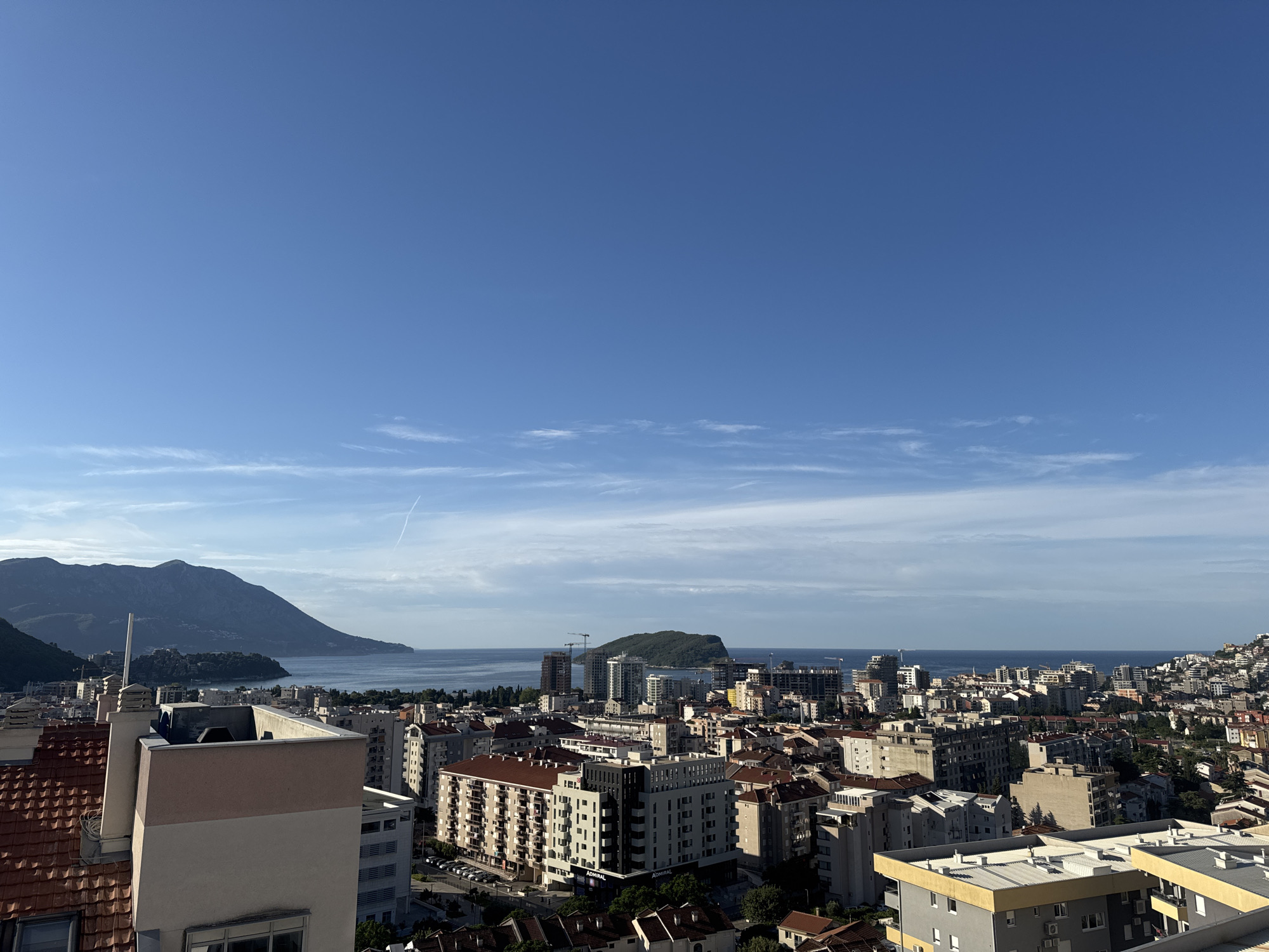 This is an aerial panoramic view of the coastal city of Budva, Montenegro, on a clear day with a bright blue sky dotted with wispy white clouds. The foreground is dominated by the rooftops of various buildings, showcasing a mix of architectural styles with terracotta tiles and modern concrete structures. Several buildings have distinct facades with visible windows and balconies.
In the mid-ground, the dense urban landscape of Budva unfolds, characterized by apartment blocks, hotels, and commercial buildings. The architecture is a blend of older, reddish-brown tiled roofs and newer, lighter-colored buildings, some of which appear to be under construction with visible cranes.
Further in the background, the Adriatic Sea stretches out, meeting the horizon. A prominent, lush green island is situated in the sea, with a few buildings visible on its shores. To the left, a large, dark, mountainous peninsula juts out into the sea, its slopes covered in vegetation. The overall scene depicts a bustling coastal city with a backdrop of natural beauty. No people or actions are discernible in this image.