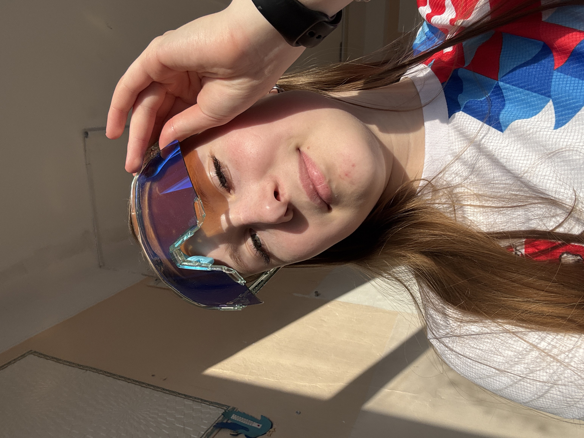 A young woman lies on her side, holding a pair of iridescent goggles near her face with her right hand. She has fair skin, long brown hair, and is wearing a white t-shirt with red and blue patterns. She is also wearing a black smart watch on her left wrist. The background appears to be an interior space with a tiled floor. The lighting suggests sunlight streaming in from a window. There is no clear indication of Usolye, Russia in this image.