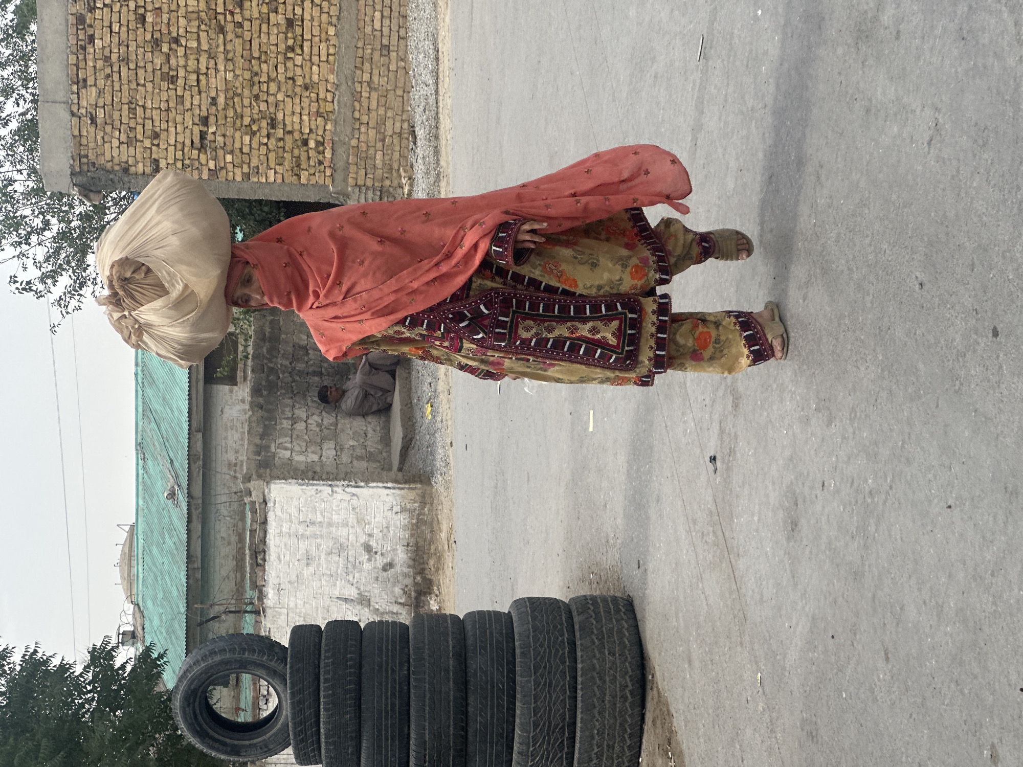In Quetta City Tehsil, Pakistan, a woman in traditional dress, headscarf, and sandals carries a large white bundle on her head. She walks on a grey paved road. Stacks of tires line the road. Behind her are buildings with brick and metal roofs. A man sits in the background.
