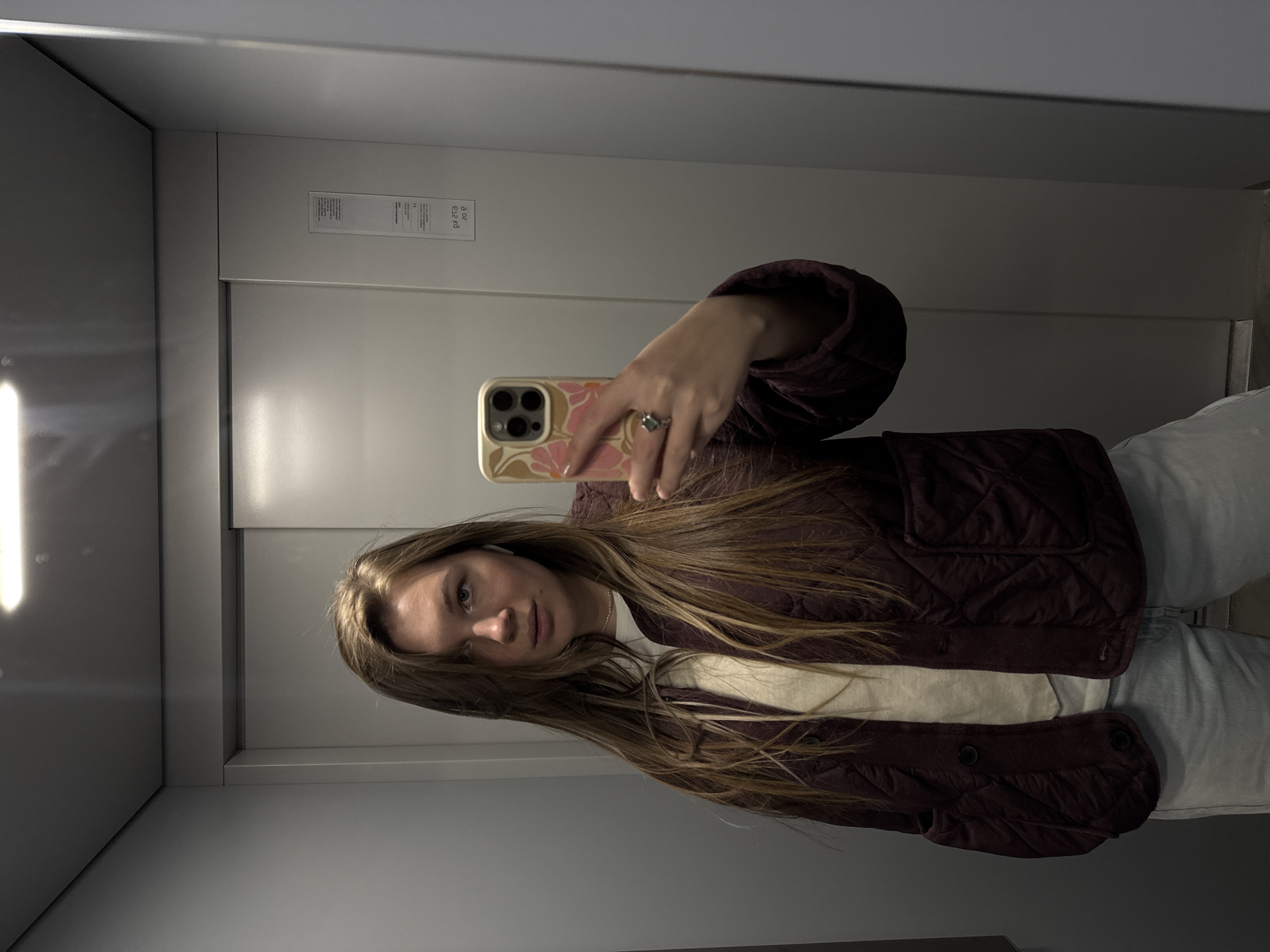 The image captures a young woman taking a mirror selfie, likely inside an elevator. She has long, straight, light brown hair and a neutral expression, looking towards the camera.
She is wearing a dark maroon or burgundy quilted jacket, open over a light-colored (possibly off-white or cream) t-shirt, and light blue jeans. On her right ring finger, she wears a silver-colored ring with a dark stone. A white earbud (AirPod) is visible in her left ear. She holds her smartphone, an iPhone with a three-camera system, in her right hand. The phone case is light-colored with a distinct pink and tan floral or abstract design.
The setting is an enclosed space with smooth, light grey or off-white panels, typical of a modern elevator interior. The image is a mirror reflection. On a white rectangular label or plaque visible on the wall, mirrored text can be deciphered as "CE", "e12 KG", and "20 C", which are likely elevator specifications or certification marks. The lighting is artificial and bright, reflecting off the metallic surfaces. The location context is Vozdovac Urban Municipality, Serbia.