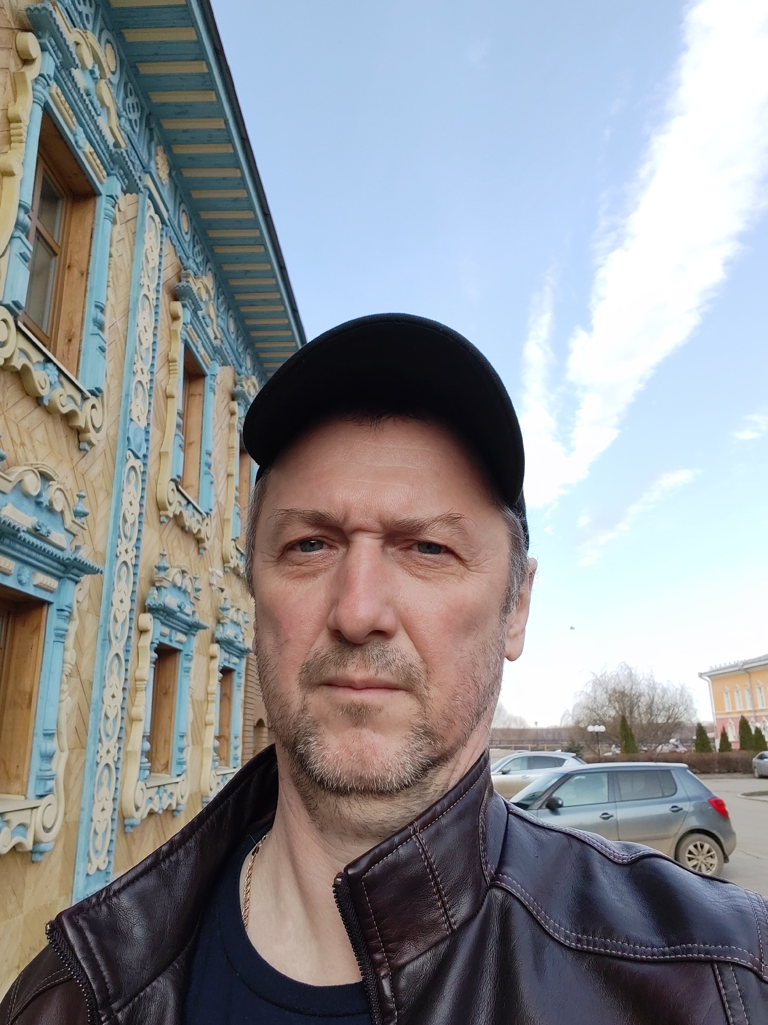 A selfie of a middle-aged man wearing a black baseball cap and a brown leather jacket. He has short hair, a stubble beard, and is looking directly at the camera.
Behind him is a two-story building with intricately carved wooden details, painted in a combination of yellow, light blue, and brown. The building has several windows.
In the background, there are parked cars, bare trees, and a building with orange columns. The sky is partially cloudy.
The location is likely Sarapul, Russia, as indicated by the architectural style of the building.