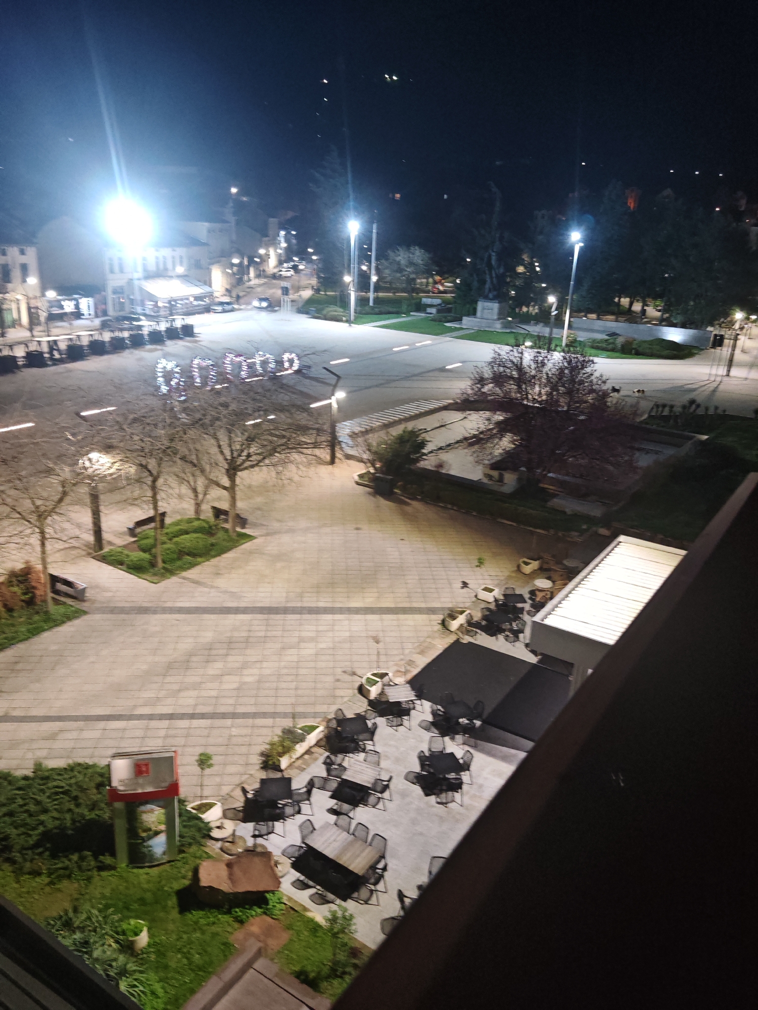 The image shows an elevated view of a town square at night. The location is the square of Hristo Botev, in the city of Vratsa, Bulgaria. Several streetlights illuminate the square, and buildings are visible in the background. The square features trees, manicured green spaces with benches, and a decorative structure spelling out the word "VRATSA" in lights. Tables and chairs, likely belonging to a cafe or restaurant, are arranged on a patio area adjacent to a building. The perspective suggests the photo was taken from an upper floor window or balcony. There is no indication of people.