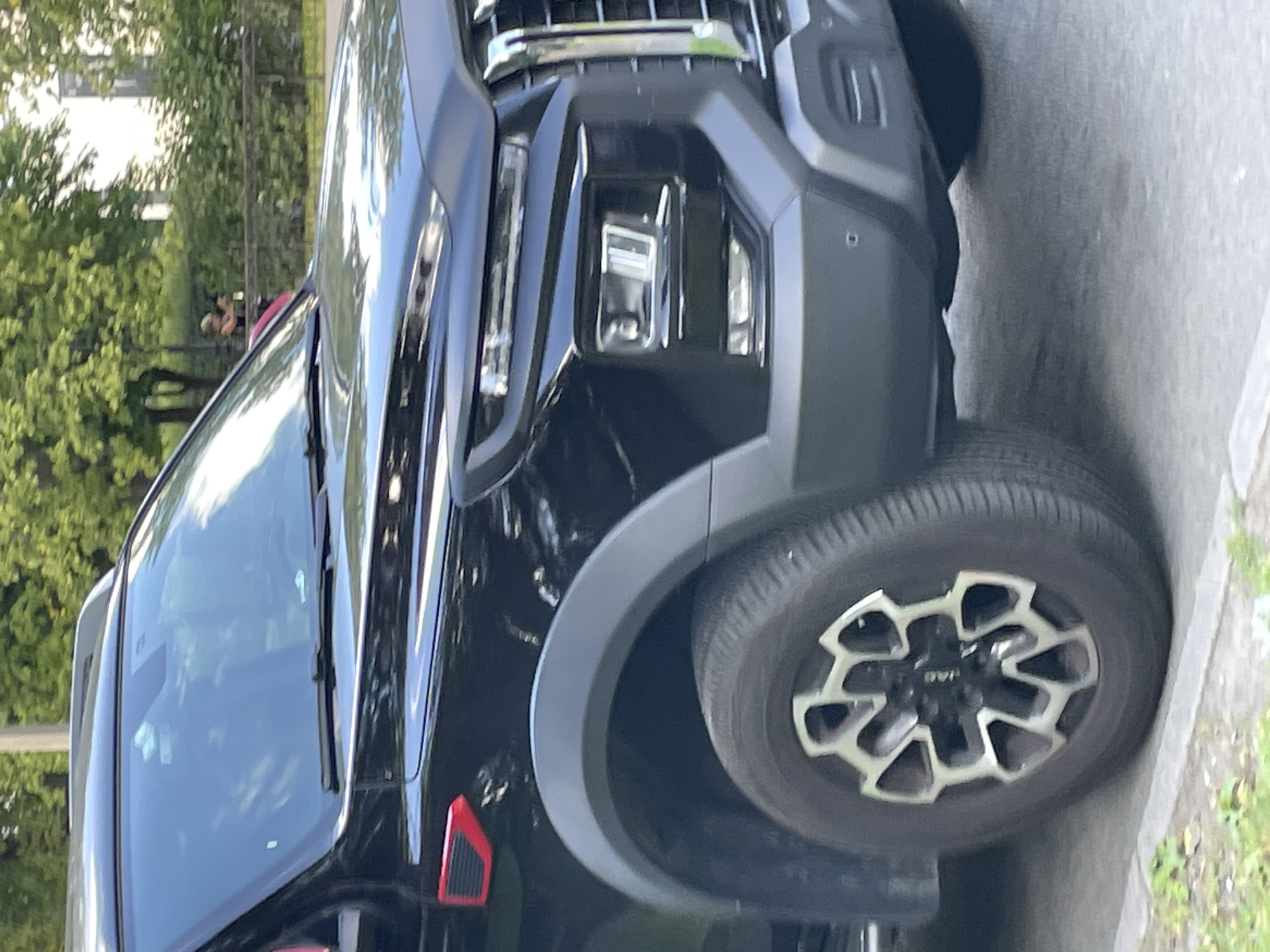 The image shows a black SUV parked on a street in Saint Petersburg, Russia. The vehicle is a modern model with a prominent grille and sharp headlights. The car has black wheel arches and a unique silver and black rim design. In the background, there are trees, a fence, and a building. A person is visible in the distance, standing near a hedge. The lighting suggests it is daytime, likely late afternoon given the shadows. The overall impression is that of a typical urban scene.