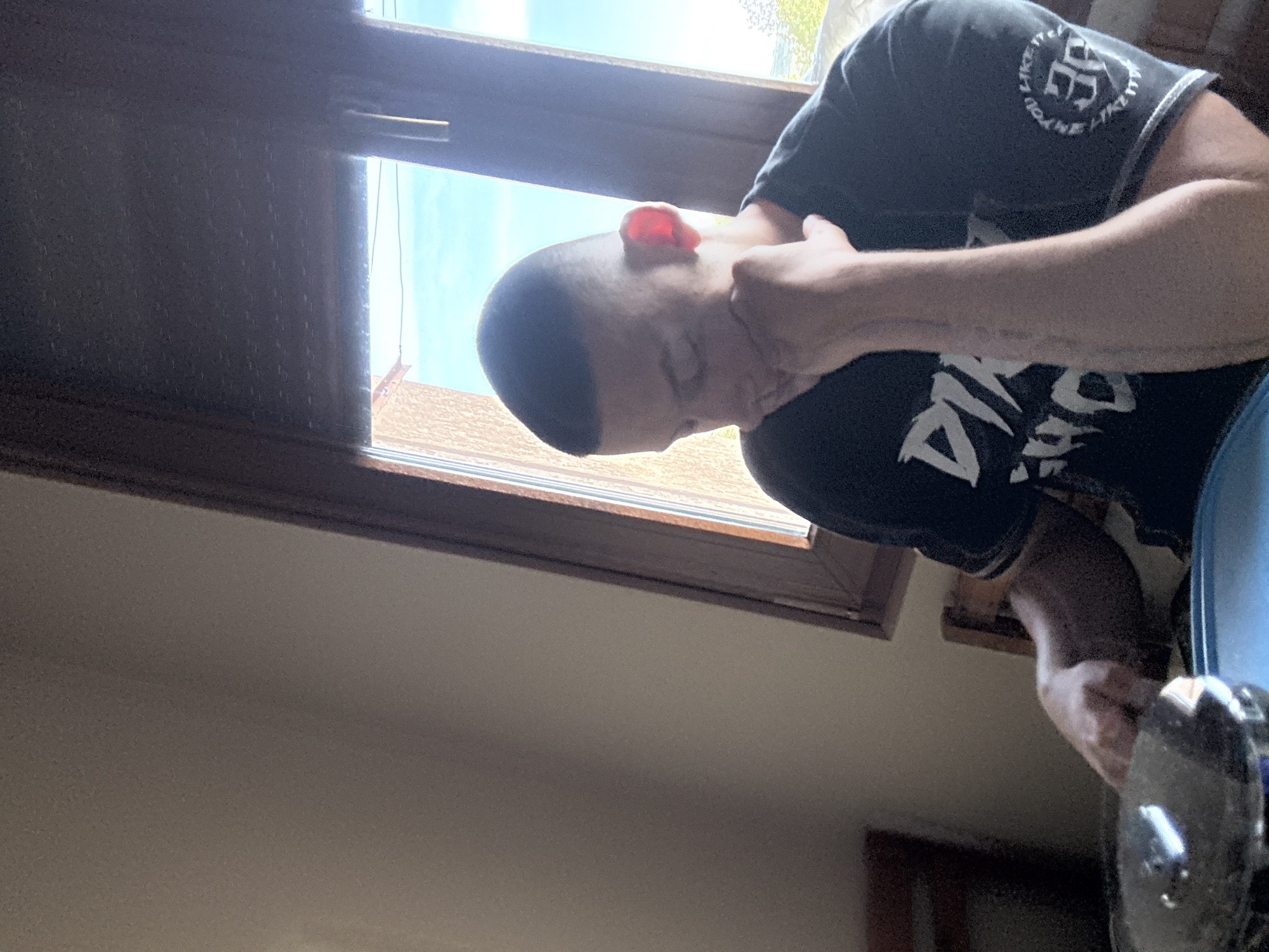 A man with a shaved head is positioned indoors, possibly in a house or apartment in Goritsa, Bulgaria. He is wearing a black t-shirt with a graphic design and a circular logo on the sleeve. The man's right hand is raised towards his neck, suggesting he might be adjusting his posture or feeling discomfort. He is holding a dumbbell in his left hand. Natural light filters through an open window behind him, revealing a glimpse of a blue sky and a section of a roof, possibly indicating a residential area in Goritsa, Bulgaria.