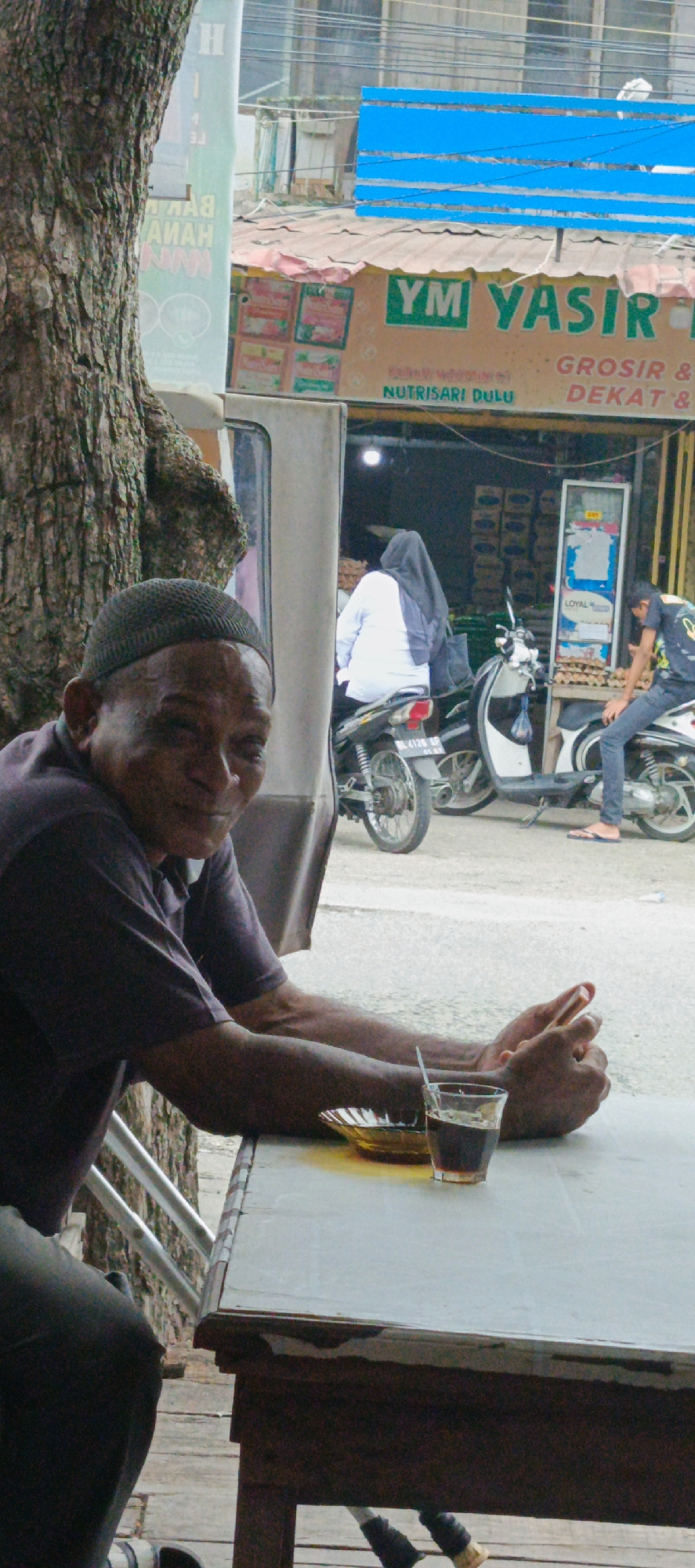 The image depicts a street scene in Garut, Indonesia, with a focus on a man sitting at a table. The man, wearing a dark purple polo shirt and a dark skullcap, is looking towards the camera with a slight smile. He is seated at a light-colored table, with his arms resting on it. A glass of dark liquid with a straw and a small amber dish are on the table in front of him. To the left, a tree trunk partially obscures the view. In the background, a storefront with a sign that reads "YM YASIR" is visible, along with motorcycles and people. One person, wearing a light blue top and a dark headscarf, is seated on a motorcycle. Another person, wearing a dark t-shirt with a graphic design, is also on a motorcycle. The storefront appears to be a shop selling goods, with stacks of items visible behind a glass display. The overall environment suggests a bustling street with commercial activity.