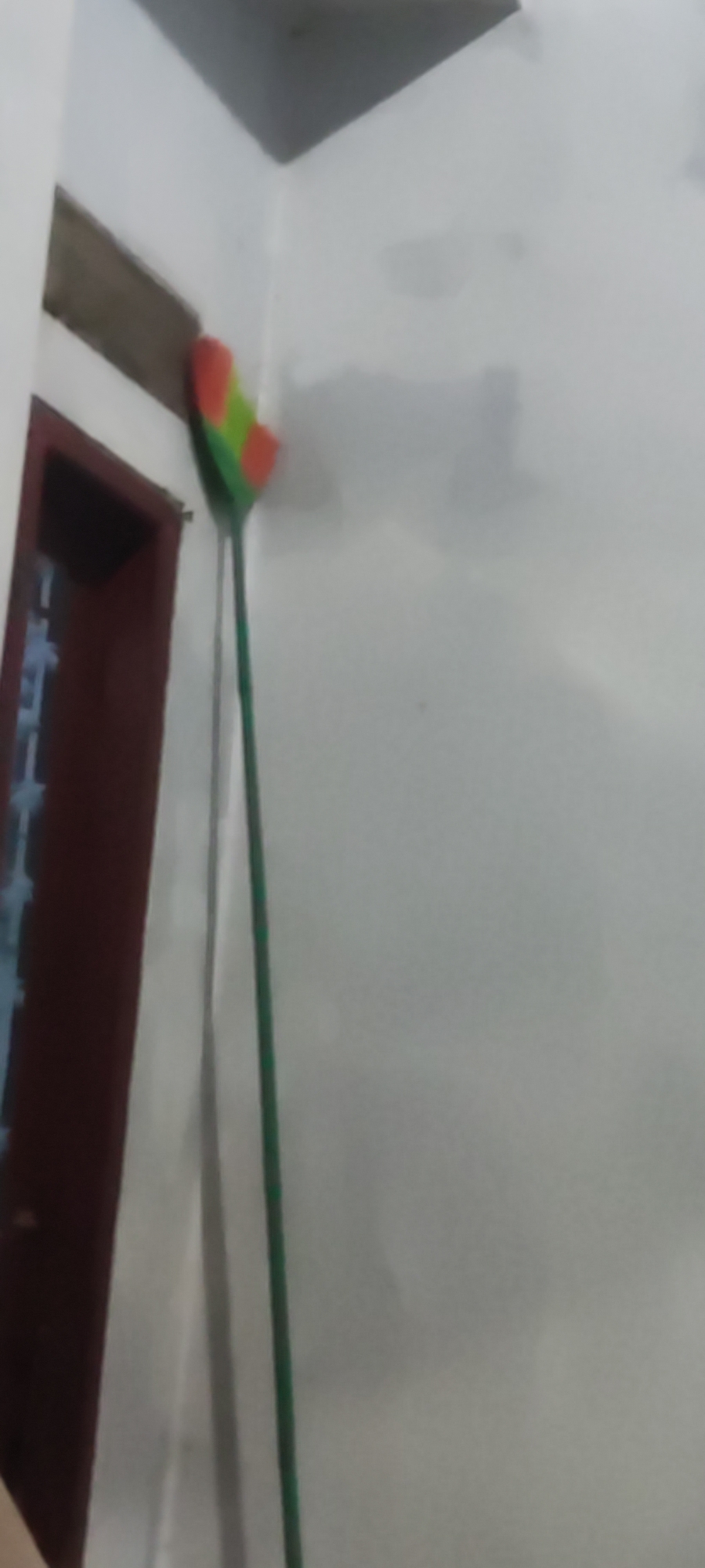 A broom with a red, yellow, and green brush head is leaning against a white wall, next to a dark brown door with a lattice design. The broom's handle is green. The scene appears to be indoors, possibly in a residential area. There is no indication of Karangwangun, Indonesia, within the image itself.