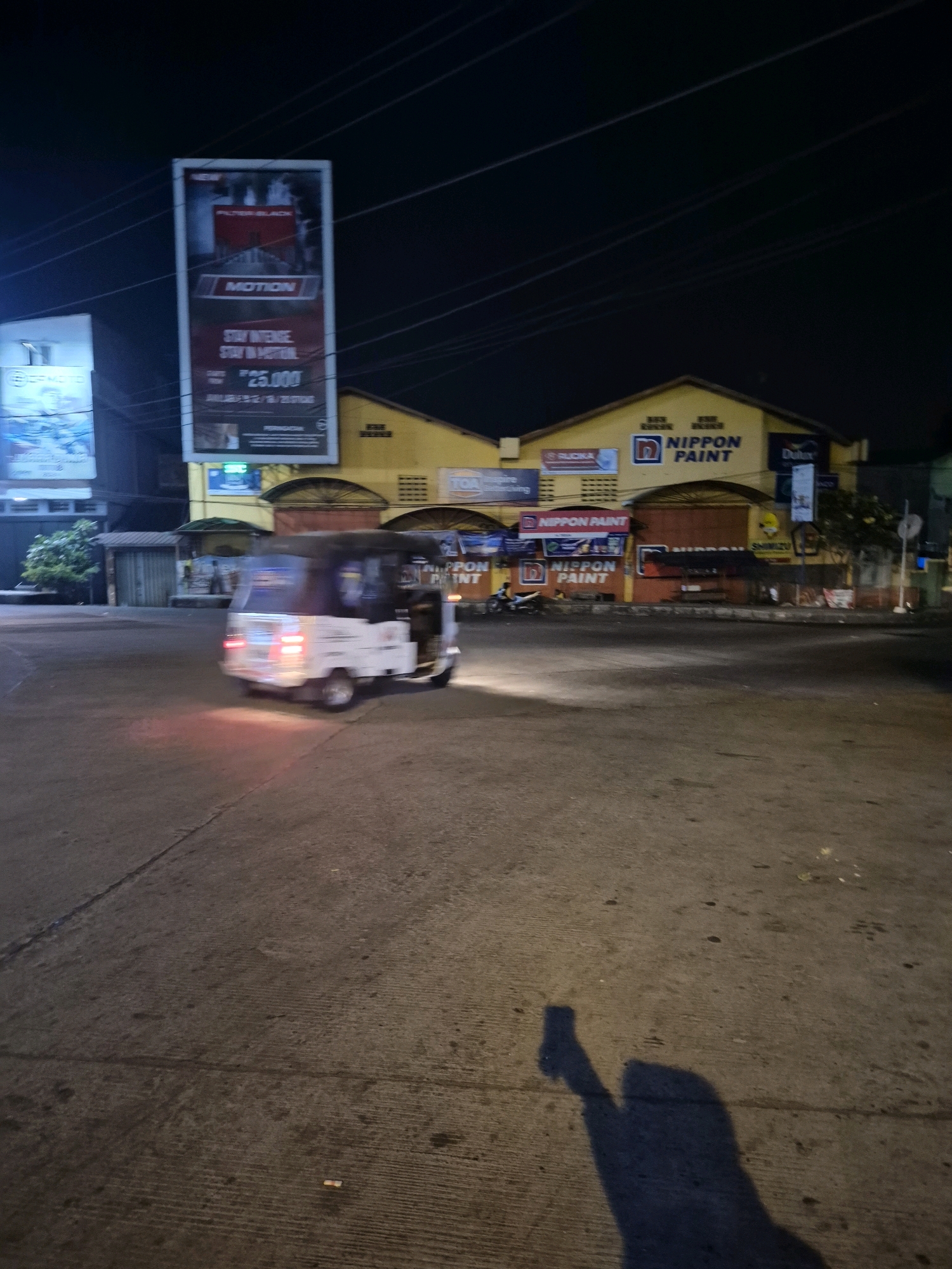 The image depicts a street scene at night in Surakarta, Indonesia. A white and black three-wheeled vehicle, commonly known as a bajaj, is moving from left to right in the foreground. Its taillights are illuminated red, indicating its movement and the low light conditions. The street appears to be paved with concrete and shows some wear and tear, with visible lines and debris.
In the background, a yellow building with a red roof dominates the scene. Several signs are visible on the building, prominently displaying "NIPPON PAINT" in large letters. Other signs for paint brands like "Dulux" and "TOA" are also present. To the left of the yellow building, a tall billboard advertising "MOTION" with text including "STAY INTENSE" and a price of "25.000" is erected. Another smaller billboard with blue and white graphics is partially visible on the far left.
The overall environment suggests an urban or semi-urban commercial area. Power lines are visible stretched across the dark night sky, crisscrossing above the buildings. The lighting is artificial, coming from streetlights and the illuminated signs. A shadow of the photographer is cast on the ground in the lower right corner of the image, with one arm raised, holding what appears to be a phone or camera.