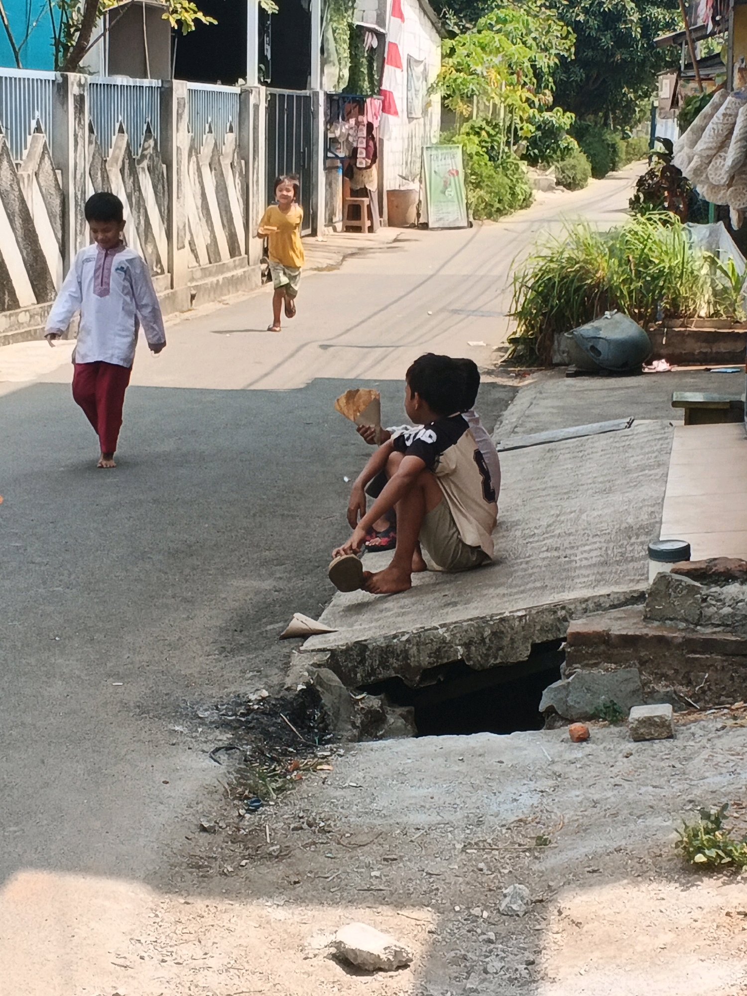 The image depicts a street scene in Kadu, Indonesia, on a sunny day. Two young boys are the central figures. In the foreground, on the right, one boy is sitting on the edge of a concrete drainage ditch, facing away from the camera. He is wearing a short-sleeved t-shirt with a design on the back and khaki shorts. He is holding a crumpled piece of brown paper in his hands. Another boy, further back and to the left, is walking away from the camera. He is wearing a white long-sleeved shirt with red trim and loose red pants, and he is barefoot. In the middle ground, a third child, also a boy, is running towards the right of the frame, wearing a yellow t-shirt and patterned shorts. The street is paved with asphalt, and to the left, a metal fence runs along the side of a property with a building. To the right, more vegetation and potted plants are visible, as well as a large grey cylindrical object. The drainage ditch in the foreground is partially collapsed and filled with debris. The overall impression is of a typical residential street in a warm climate.