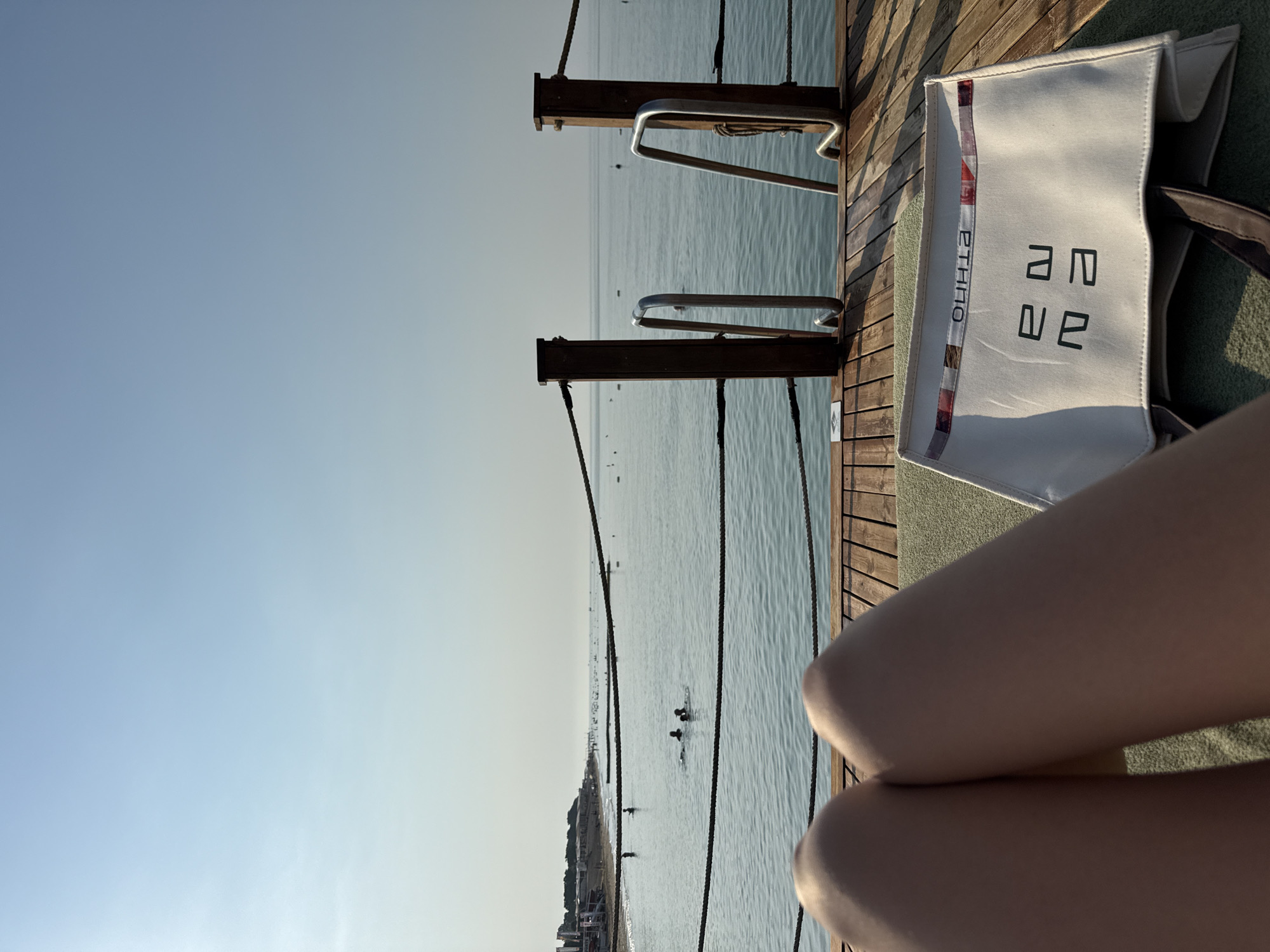 The image depicts a serene beach scene on a clear day, likely in Serik, Turkey. In the foreground, a person's legs are visible, resting on a sun lounger. In front of the lounger is a white canvas tote bag with dark brown straps, displaying an abstract pattern along the top edge and a logo or design in black lettering on its front. The lounger itself is positioned on a wooden pier that extends over the calm, blue water. The pier is equipped with wooden posts and metal railings, leading down to the water, suggesting a place for swimming or diving. In the middle ground, several people are seen swimming in the sea. Beyond them, the coastline is lined with a sandy beach, dotted with beach umbrellas and buildings, indicating a resort or populated area. The horizon is marked by a hazy line separating the sea from the pale blue sky, which dominates the upper portion of the image. The overall atmosphere is one of relaxation and leisure by the sea.