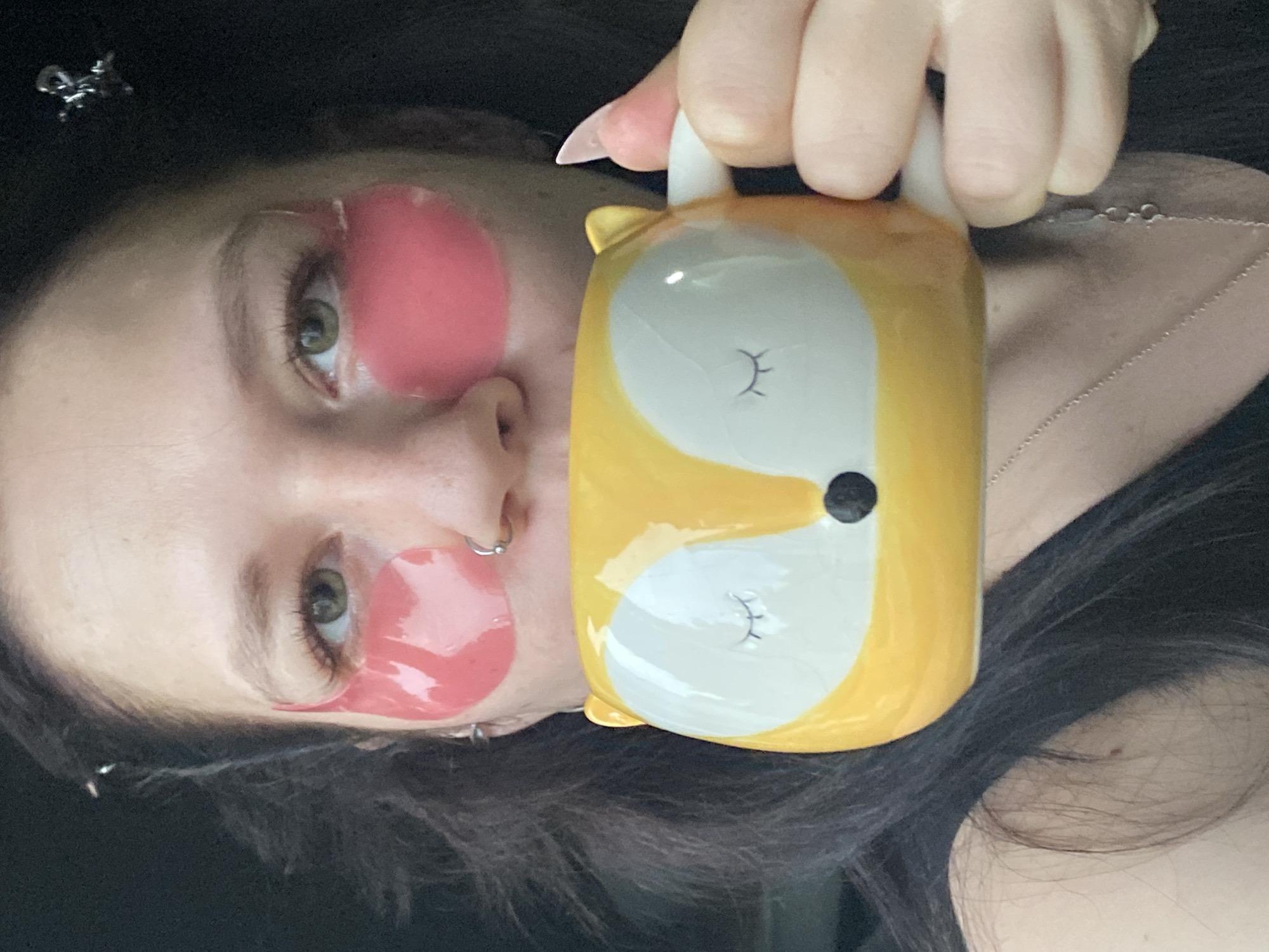 A woman is holding a fox-shaped mug to her face, obscuring part of it. She is wearing pink eye patches under both eyes and a silver nose ring. She has dark hair and a silver necklace. The background is dark and indistinct. There is no indication of the location being the city of Chaikovsky, Russia.
