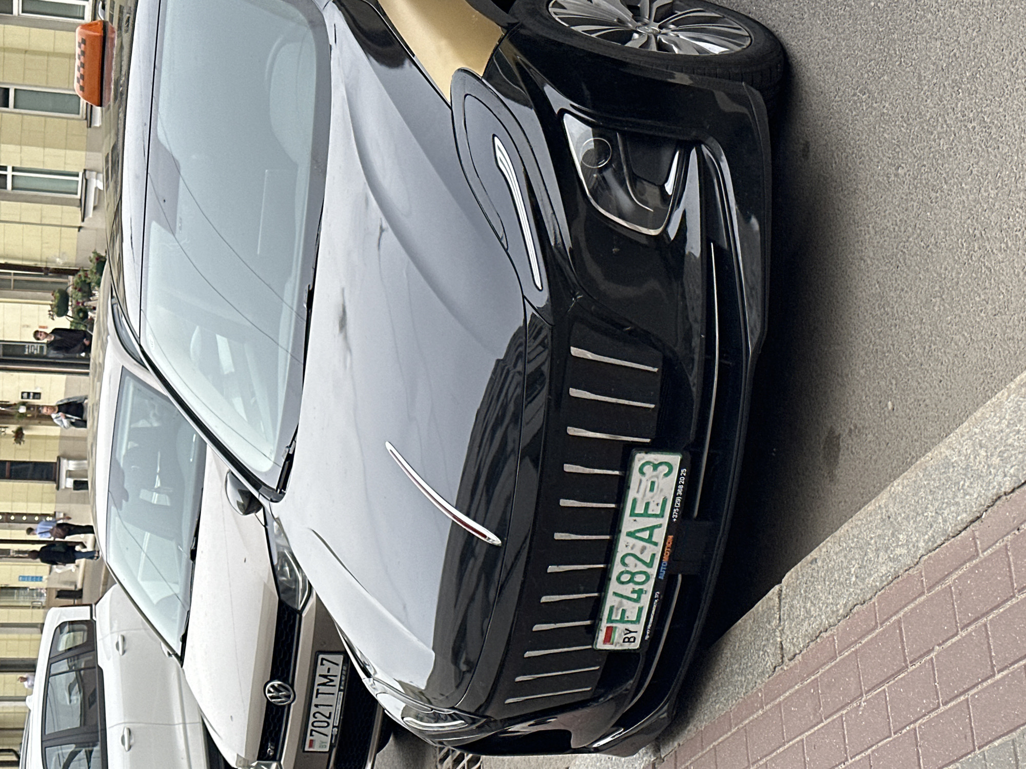 This image captures a street scene in Minsk, Belarus, featuring a black and gold taxi prominently parked in the foreground. The vehicle is a Geely Geometry C, identified by its distinctive front grille composed of multiple vertical silver bars, elongated, curved LED daytime running lights, and a central chrome stripe with a red inset running vertically down the hood. An orange taxi light is affixed to the vehicle's roof. Its Belarusian license plate, "BY E482 AE-3," is mounted below the grille, with the text "AUTOMOTION" and a Belarusian phone number, "+375 (29) 368 20 25," visible beneath it. The taxi is parked adjacent to a brick-paved sidewalk with a grey curb.
Behind the black taxi, a beige Volkswagen Polo is partially visible, displaying a Belarusian license plate reading "7021 TM-7." Further to the left and in the background, a white SUV or minivan is also partially visible.
The background shows an urban environment with several people walking on the sidewalk. Two individuals are walking together in the middle distance towards the left, while another person in a dark jacket is walking away from the camera on the far left. A man in a light jacket stands on the right side of the street. Buildings with multiple windows and classical urban architecture, including one with a yellow facade, line the street. The street surface is asphalt.