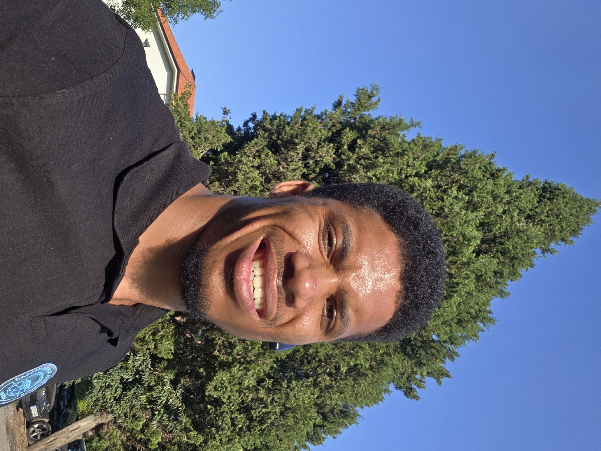 A man with dark skin and short, dark, curly hair is pictured from the chest up, smiling broadly at the camera. He is wearing a black polo shirt with a small, circular blue and white logo on the left side of his chest. He has a light beard and a faint mole on his nose. Behind him, a tall, green evergreen tree dominates the background, with a blue sky visible above. To the left of the tree, a white building with a red-tiled roof is partially visible. The scene appears to be outdoors, likely in a residential area. The man's attire and the background suggest a sunny day. The logo on the shirt contains text, including "VARNA".