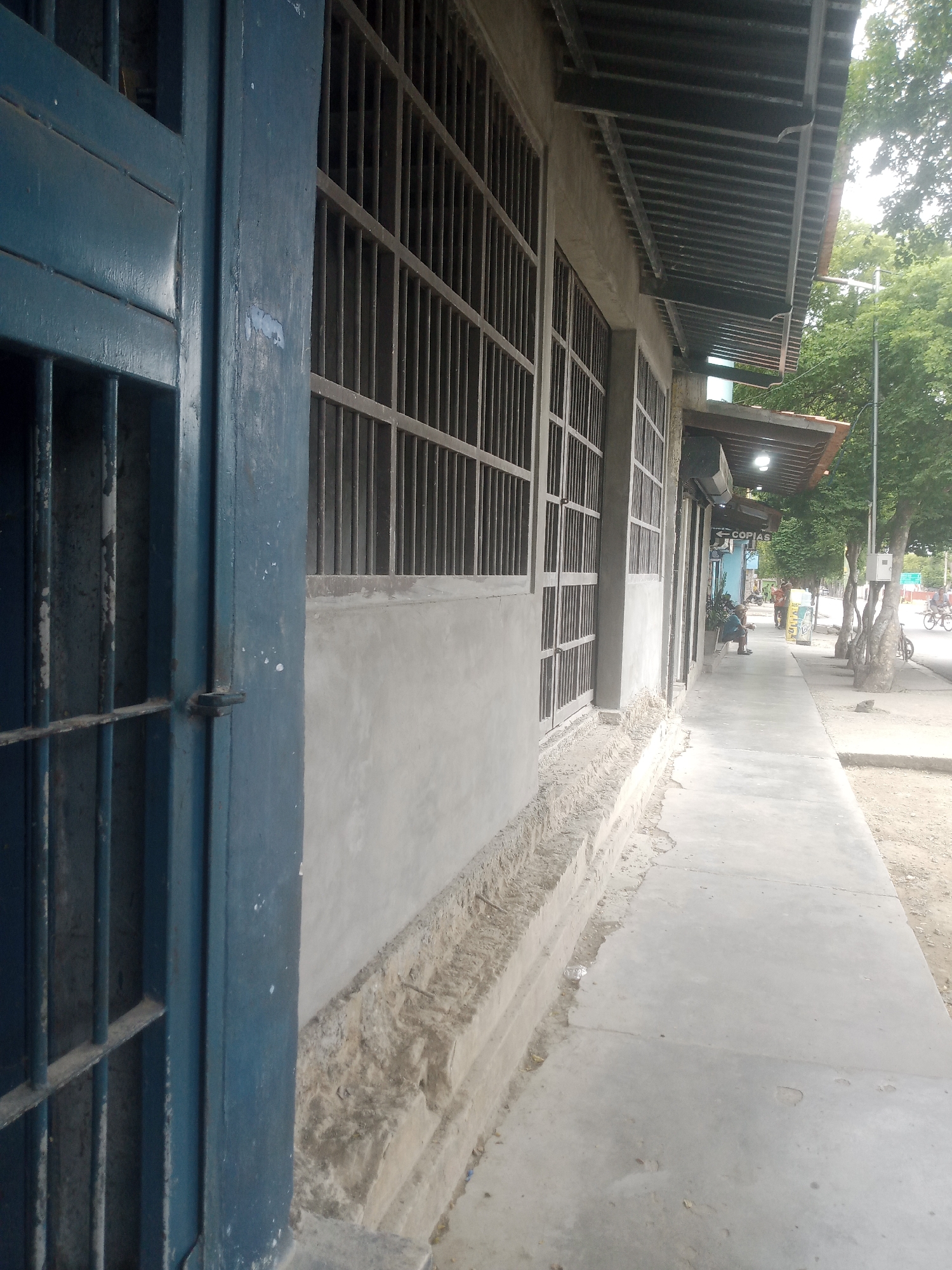 The image depicts a building with barred windows and a blue metal gate on the left. The building appears to be a commercial establishment with a sidewalk in front. In the background, trees line the street, and a few people and bicycles are visible. The scene suggests a street in a populated area, possibly a town or city. The presence of barred windows might imply a security concern or a specific type of establishment. The overall impression is that of a functional urban environment. The location is Santa Ana, Venezuela.