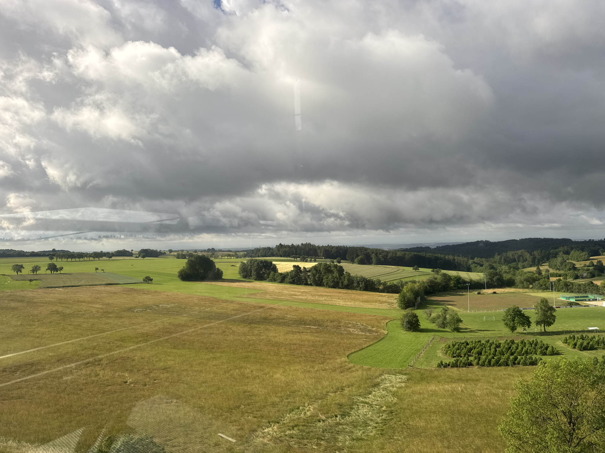 This is an aerial, wide-angle, landscape photograph of rolling green fields and trees under a dramatic sky with heavy, grey clouds. The sun is breaking through in a few spots, casting light on sections of the landscape. In the foreground, there are large fields of dry, golden grass with distinct linear patterns, possibly from harvesting. Further back, the fields transition to vibrant green pastures and cultivated areas. Scattered trees dot the landscape, and a small cluster of young evergreen trees are visible in neat rows. In the middle ground, a sports field with goalposts and what appears to be a fenced-off area with bleachers or seating are present. The background consists of a dense line of trees forming a dark horizon, beyond which lie rolling hills fading into the distance. The scene appears to be rural and agricultural, consistent with the location of Strümpfelbrunn, Germany, a village known for its scenic countryside. There are no people or animals visible in the image. The overall impression is one of natural beauty and open space.
