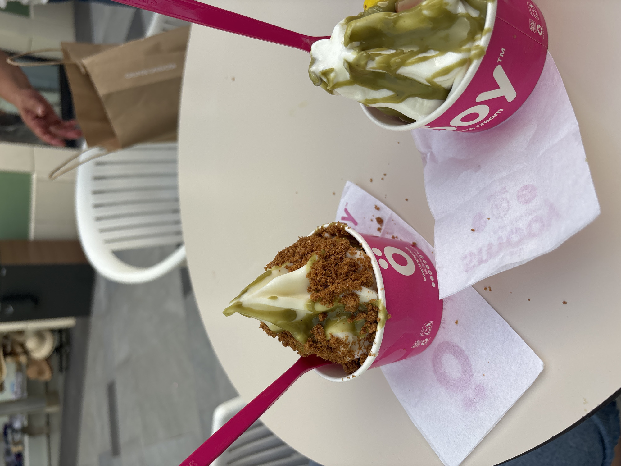 In Orihuela, Spain, at a table outside, two cups of soft-serve ice cream are displayed. The cups are pink and branded with "O YOY". The ice cream in the left cup is topped with crumbled cookies and a green sauce, with a pink spoon sticking out. The ice cream in the right cup is also swirled and drizzled with the same green sauce, with a portion of a yellow fruit visible at the base. White napkins with a pink logo are placed beneath each cup. In the background, a white plastic chair and a brown paper bag are visible, suggesting a retail or cafe environment. The table is light-colored.