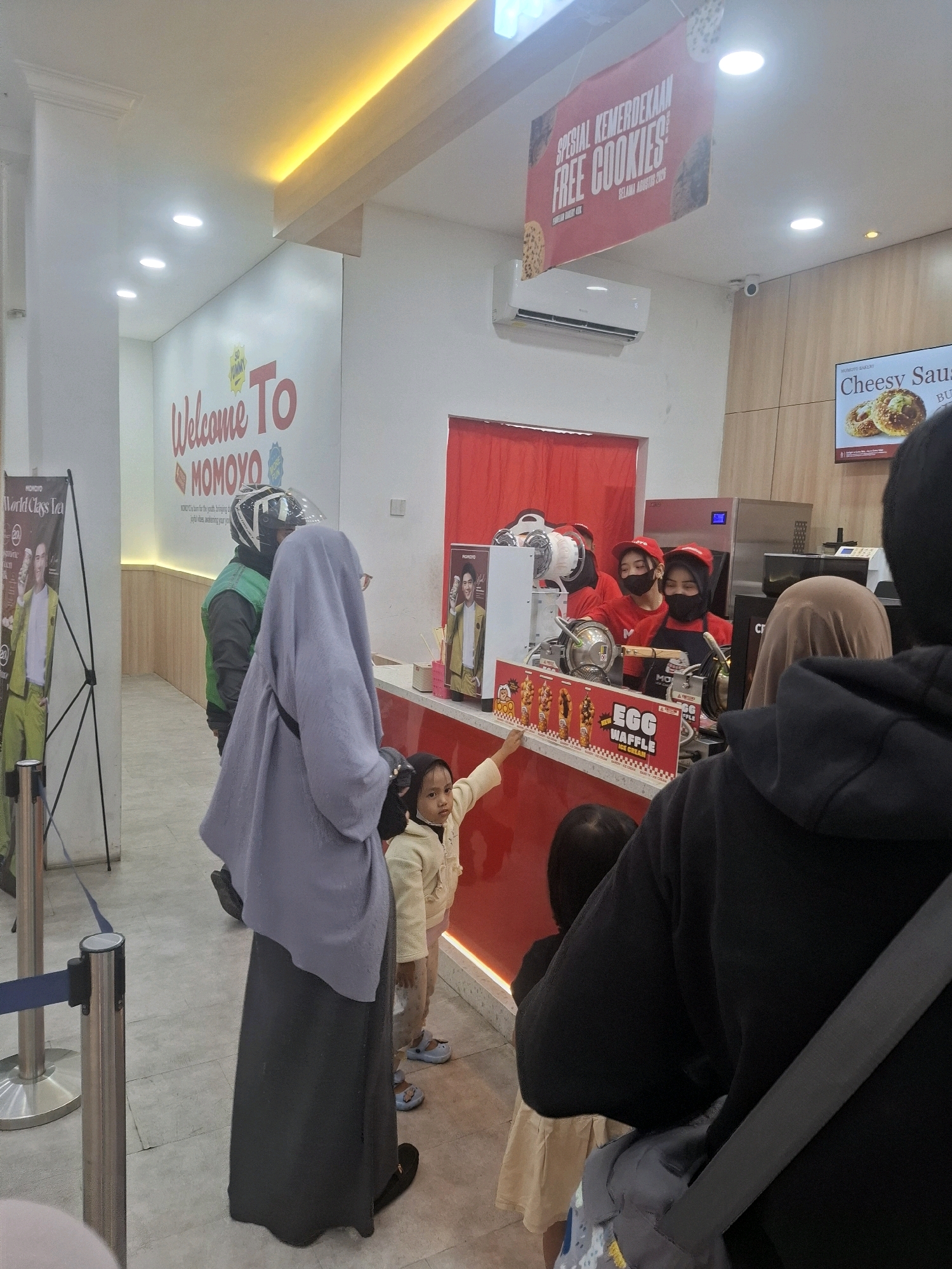 The image depicts the interior of a Momoyo store, likely a fast-food or dessert shop, located in Cianjur, Indonesia. The store features a bright, modern aesthetic with white walls and ceiling, accented by yellow LED lighting. A prominent "Welcome To MOMOYO" sign is visible on the back wall. In the foreground, a queue of people are waiting to be served at a counter where employees, wearing red shirts and head coverings, prepare food items such as "Egg Waffles." A digital menu board displays "Cheesy Sausage." The atmosphere appears to be busy, with customers and staff interacting within the commercial space. The overall scene captures a typical retail environment in a bustling Indonesian city.