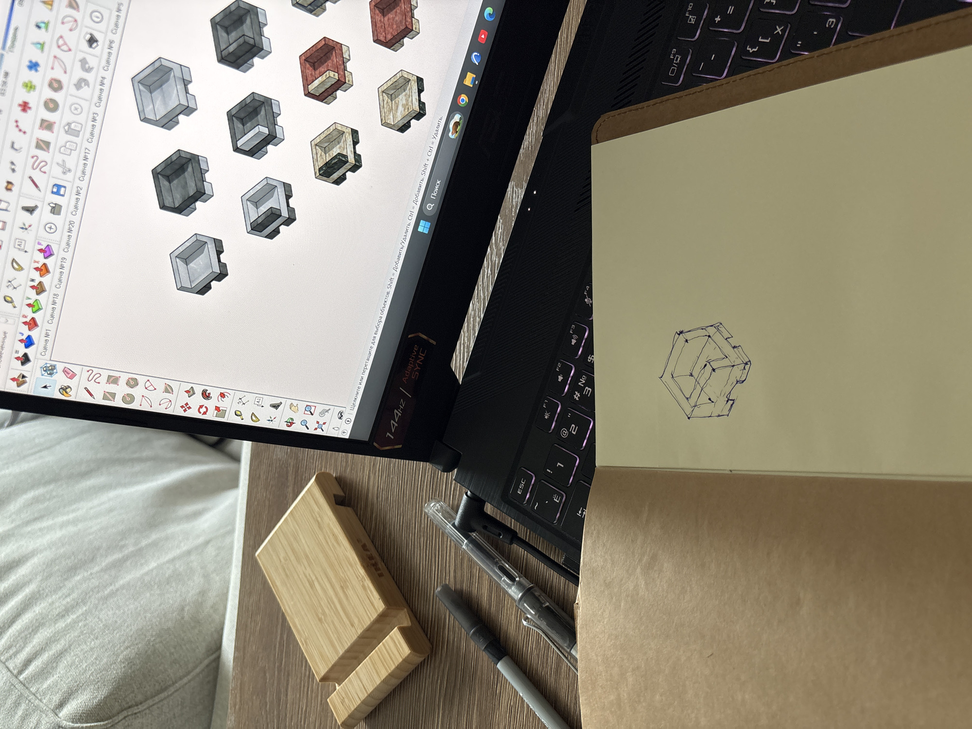 The image shows a workspace on a table. A laptop is open, displaying a 3D modeling program with several cube-like designs. A notebook is placed next to the laptop with a pen drawing of one of the cube designs. A wooden phone stand and two pens rest on the table. The environment appears to be indoors, with part of a cushioned surface visible. The image doesn't provide enough information to determine any location in Thailand.