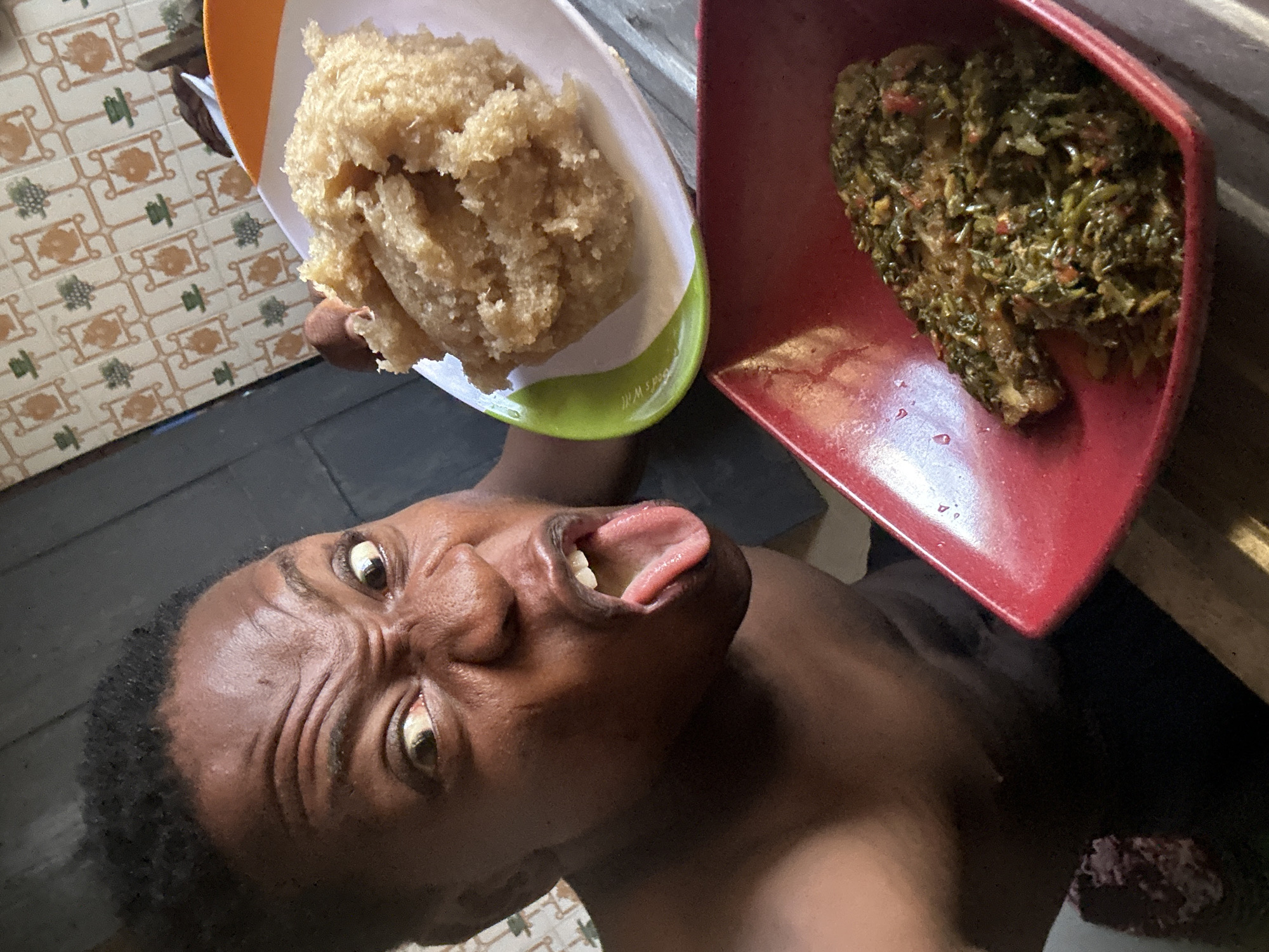 The photograph shows an individual holding two dishes of food. The person is of African descent and has a furrowed brow, wide eyes, and an open mouth with the tongue protruding. One dish, held in the left hand, contains a mound of beige-colored food with a coarse texture. The second dish, held in the right hand, is filled with a dark green leafy vegetable preparation.
The individual is positioned indoors. The floor is tiled with an intricate brown and beige pattern. A grey floor and a wooden trim are also visible. The location in the city is Unknown, Unknown.