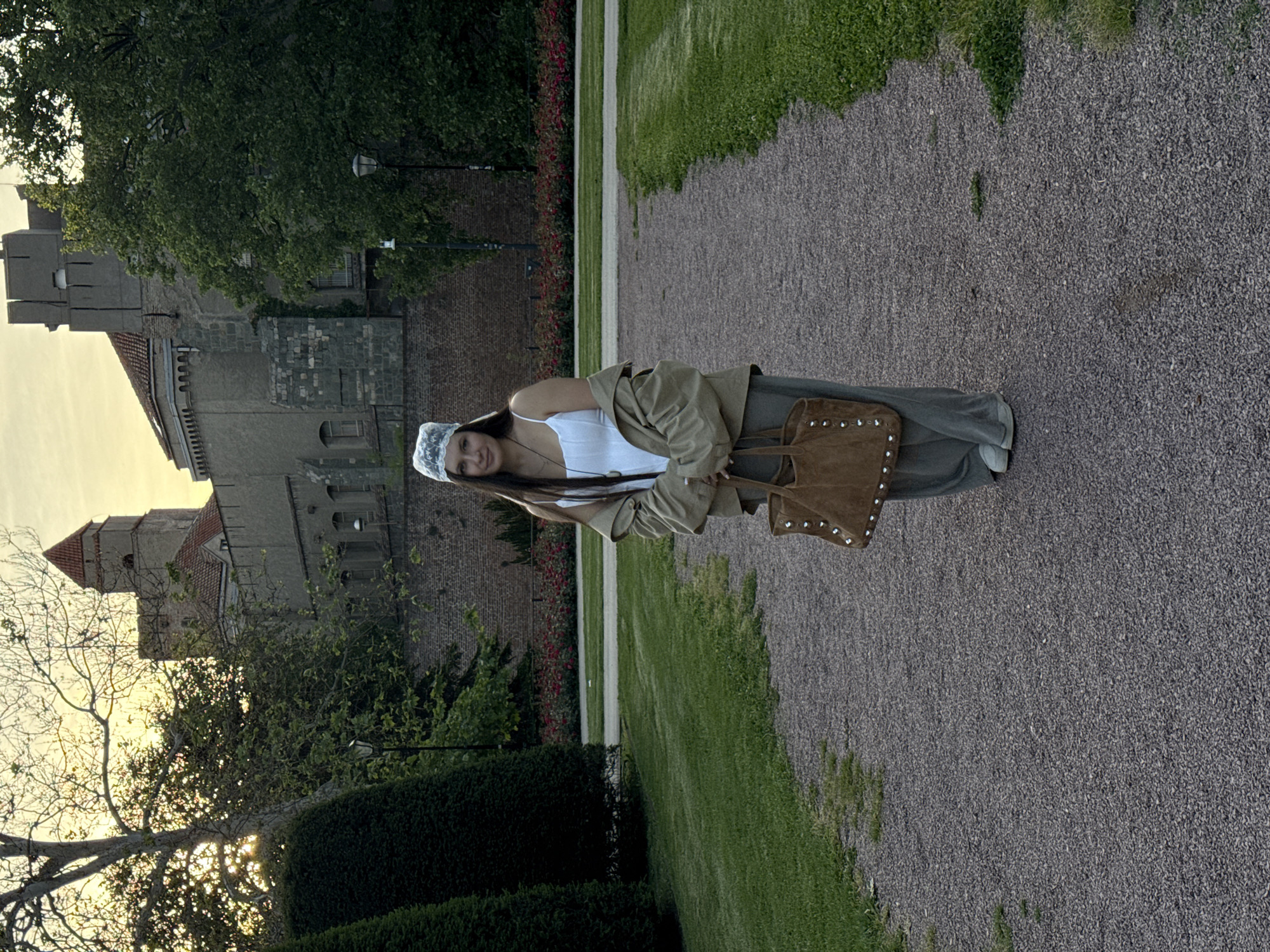 A woman in a white top, khaki pants, and a tan jacket, stands on a gravel path in front of a castle. She wears a white bonnet and holds a tan tote bag. The castle is made of grey stone with a red tile roof. Green grass and bushes surround the path and castle. The sky is golden. The location is likely Kalemegdan Park in Belgrade, Serbia.