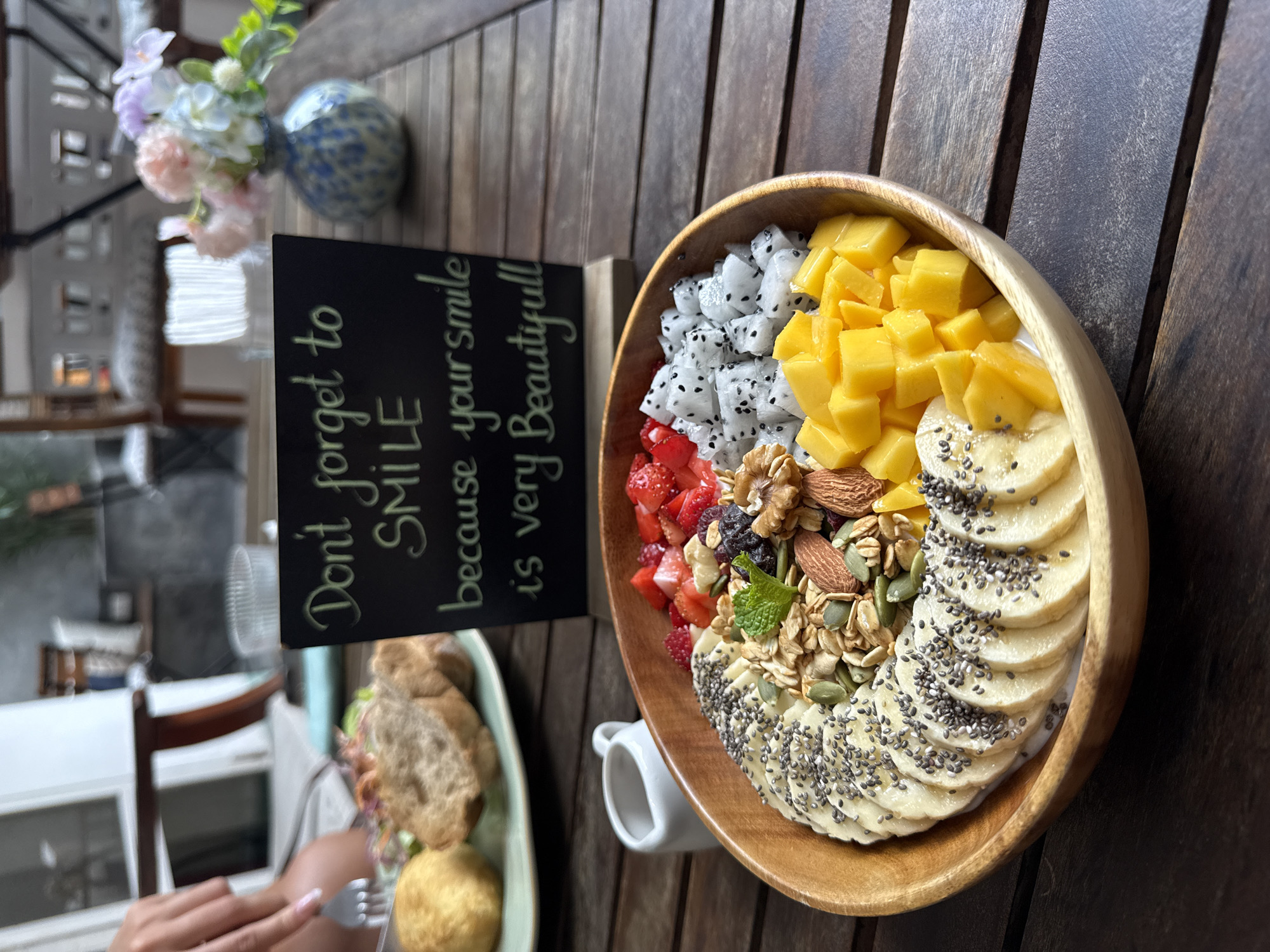 The image captures a wooden table setting in Đà Nẵng, Vietnam. A wooden bowl is filled with a colorful breakfast or brunch dish consisting of dragon fruit, diced mango, strawberries, nuts, granola, sliced bananas sprinkled with chia seeds, and a white liquid base, presumably yogurt or a smoothie. Next to the bowl stands a small chalkboard with the message "Don't forget to SMILE because your smile is very Beautiful!" In the foreground, a hand is holding a fork next to a green plate with a sandwich and a fried item. The setting appears to be outdoors, as evidenced by the natural light, the plant on the table, and the partial view of a building in the background. A small white pitcher is also visible on the table. The overall atmosphere suggests a casual, health-conscious dining experience.