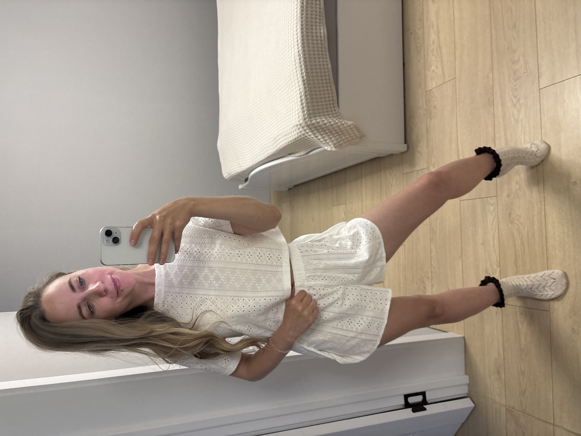 A young woman is taking a selfie in what appears to be a bedroom. She is wearing a matching white eyelet top and shorts with patterned socks with a black ruffled edge. She is holding a light-colored smartphone. The room contains a bed with a white textured cover, light wooden flooring, and white walls. There is no information provided in the image to confirm that this is in Chaikovsky, Russia.