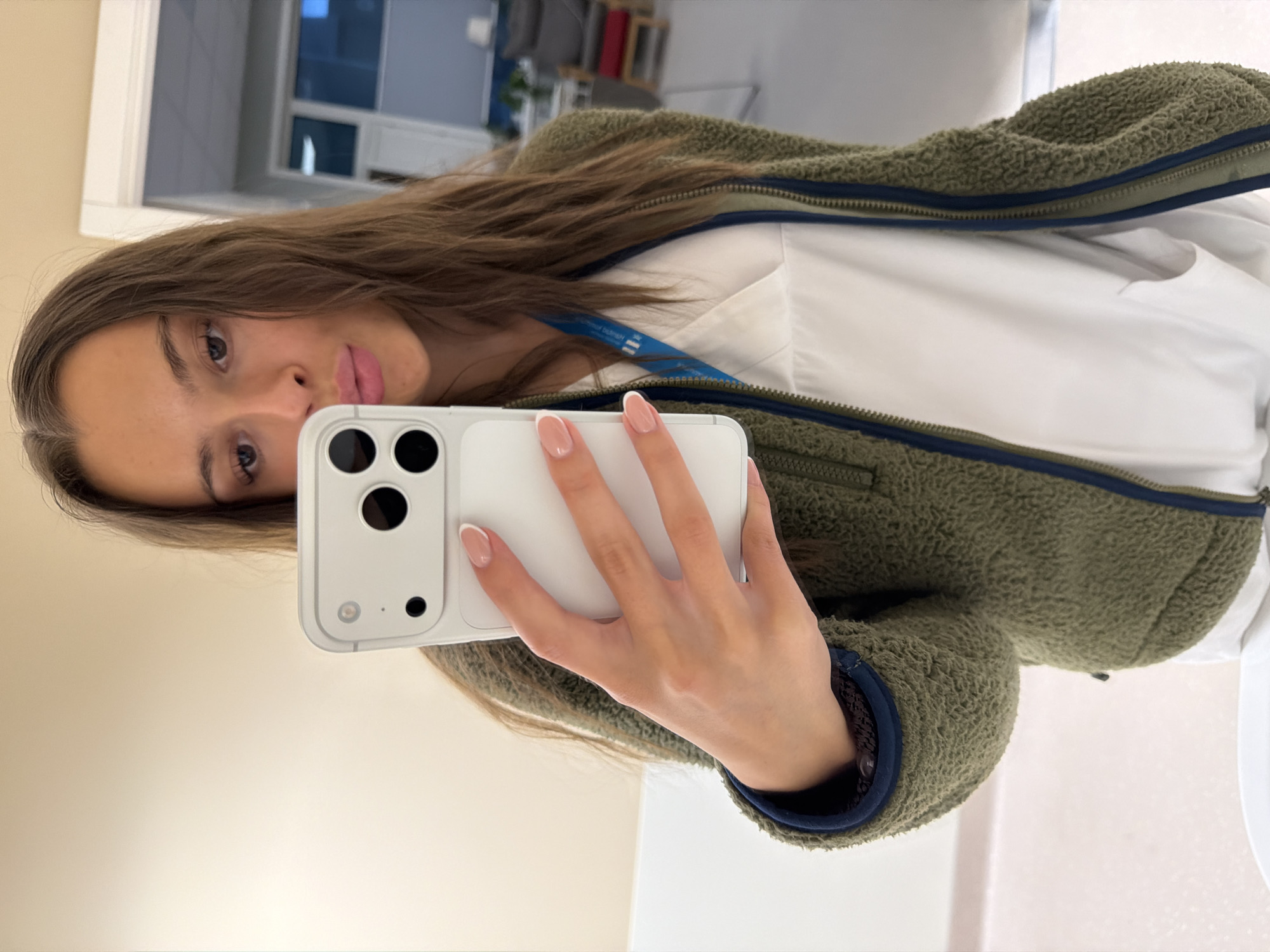 A young woman with long, brown hair is taking a selfie in a mirror. She is wearing a white scrub top and an olive green fuzzy jacket. She is holding a white iPhone in her right hand, which is angled to capture her reflection. Her left hand is resting on the phone. The background appears to be an indoor setting with beige walls and a doorway leading to a room with windows. The lighting is bright and even. The woman is looking directly at the camera with her lips slightly parted. This scene is likely taking place within the Harstad Hospital (Sykehuset i Harstad) in Harstad, Norway, given the presence of medical scrubs and the professional environment suggested by the background.