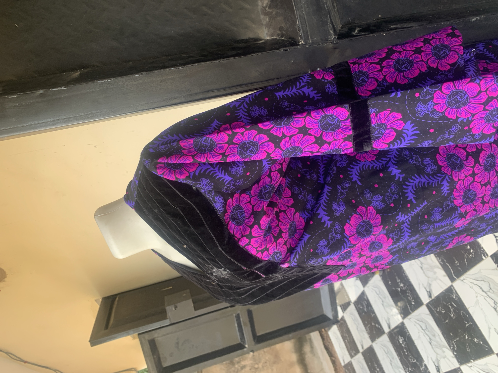 A headless mannequin displaying a black garment with a vibrant floral pattern of pink and purple flowers is positioned outdoors in front of a dark doorway. The garment features black velvet trim along the neckline and cuffs. The mannequin is on a black and white checkered floor. Behind the mannequin, a light-colored wall and a portion of a doorway are visible. The overall setting appears to be an exterior, possibly a storefront or an outdoor display area, in Jauro Yinu, Nigeria.