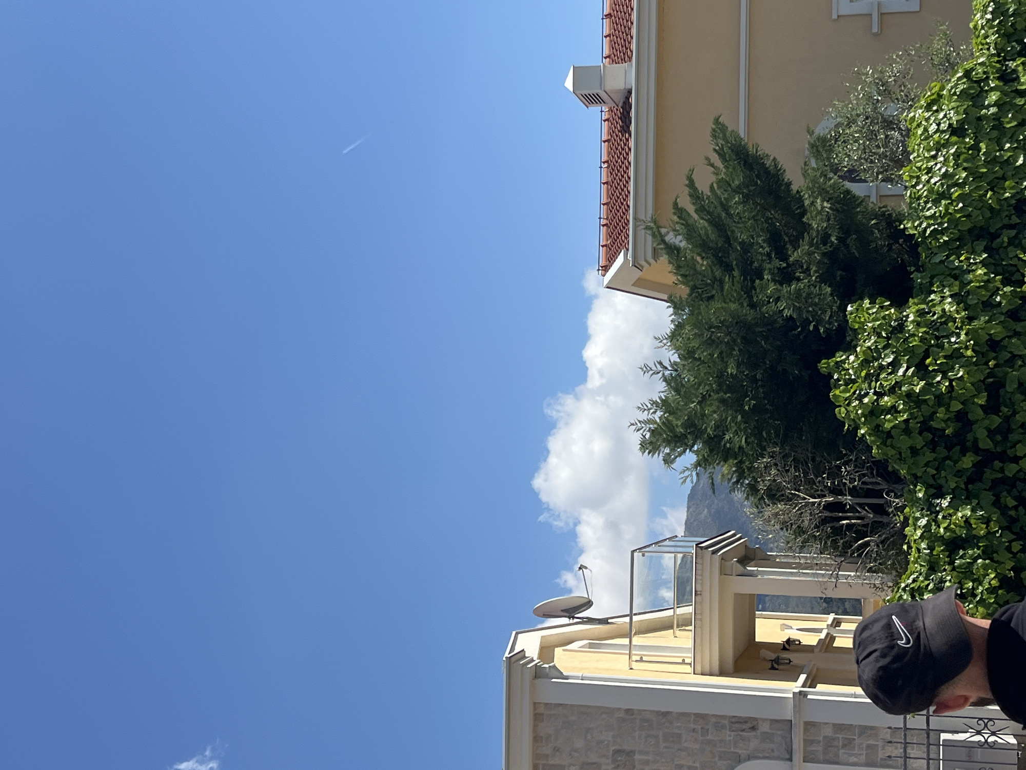 The image shows a scene in Boreti, Montenegro, focusing on the architectural details of a building and the natural environment. A man with a black cap is partially visible in the lower right corner. A building with a red-tiled roof, yellow walls, and white trim is partially visible. Green trees and lush foliage cover part of the building. The upper left portion of the image is filled with a clear blue sky and a faint airplane contrail. Part of a building with a stone facade and satellite dish is visible, with a glass enclosure on the roof. A white cloud is behind the satellite dish.