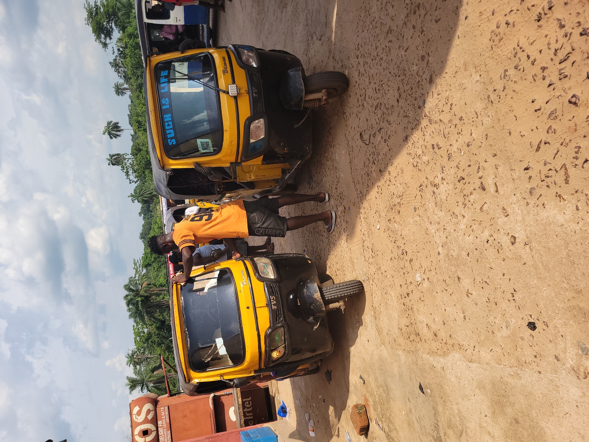 In the city of Egbagu, Nigeria, under a cloudy sky, two yellow auto-rickshaws, also known as Keke Napeps, are parked on a dirt and concrete surface. The auto-rickshaws are the central subjects of the image. The one on the left is parked in front of a red and orange kiosk with the logo "OS" visible. The kiosk has "DELIGHT FIN RECHARGE CARDS & DATA" printed on it, along with phone numbers. A portion of an Airtel logo is also visible on the kiosk. The auto-rickshaw on the right has the phrase "SUCH IS LIFE" written in blue lettering on its windshield.
A man wearing an orange t-shirt with the number "9" on it, dark shorts, and sandals stands next to the auto-rickshaw on the left, appearing to be engaged in a conversation or transaction. Another person, wearing a pink shirt, is visible inside the auto-rickshaw on the right. The environment appears to be a roadside market or transportation hub, with other vehicles and debris visible in the background, including what appears to be a white auto-rickshaw with a blue stripe. Palm trees and other lush green vegetation are seen in the distance, indicating a tropical climate. The ground is a mix of loose dirt, gravel, and patches of concrete, with some scattered litter. Sunlight casts shadows on the ground, suggesting it is daytime.