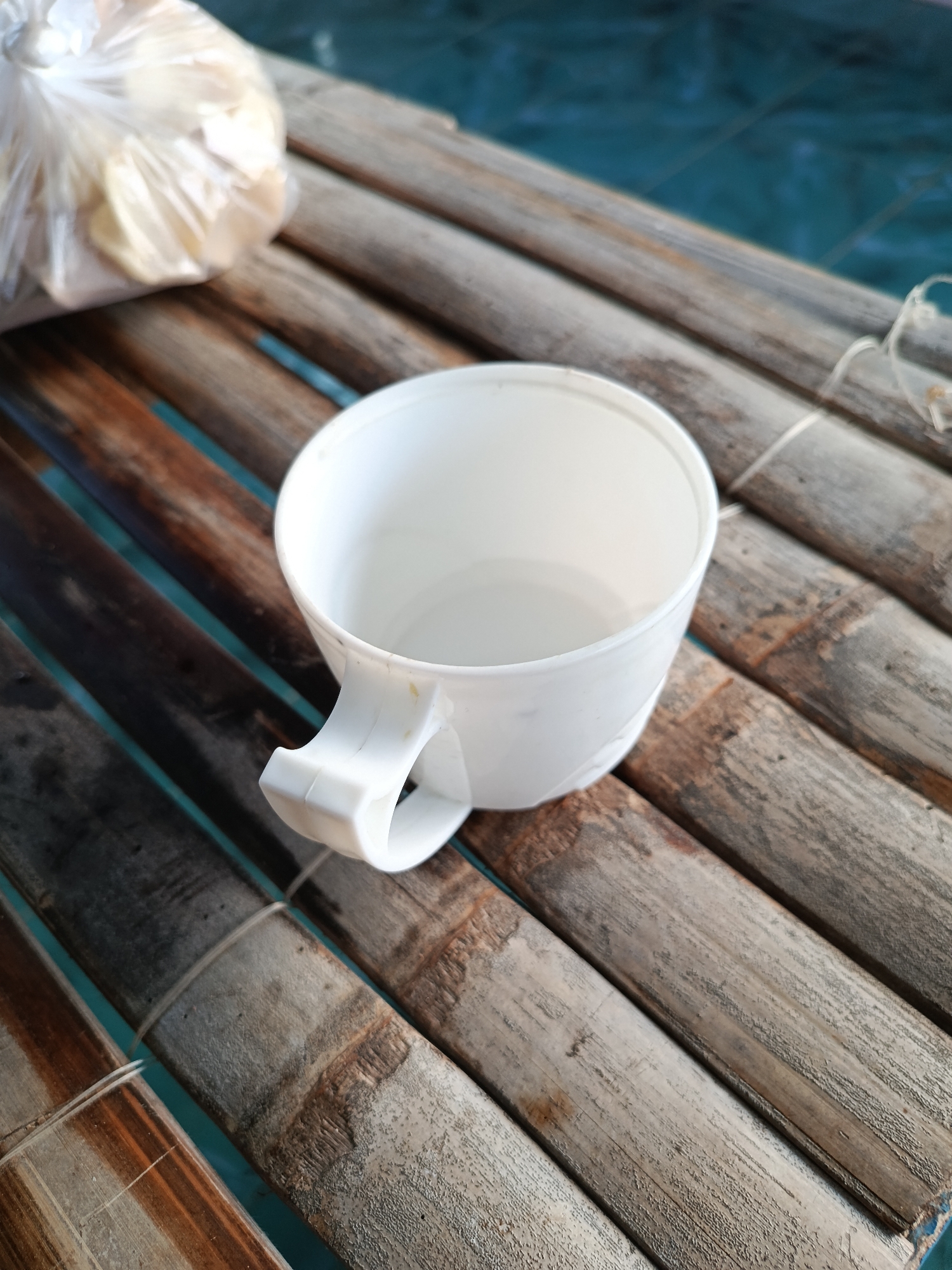 In the city of Mansalean, Indonesia, on a surface composed of parallel bamboo poles, a white plastic mug with a handle is placed. A plastic bag filled with a light-colored substance is visible to the left of the mug, partially obscuring some of the bamboo. The bamboo poles are laid out horizontally across the frame, with some appearing weathered and worn. The background consists of a blue surface, possibly water.