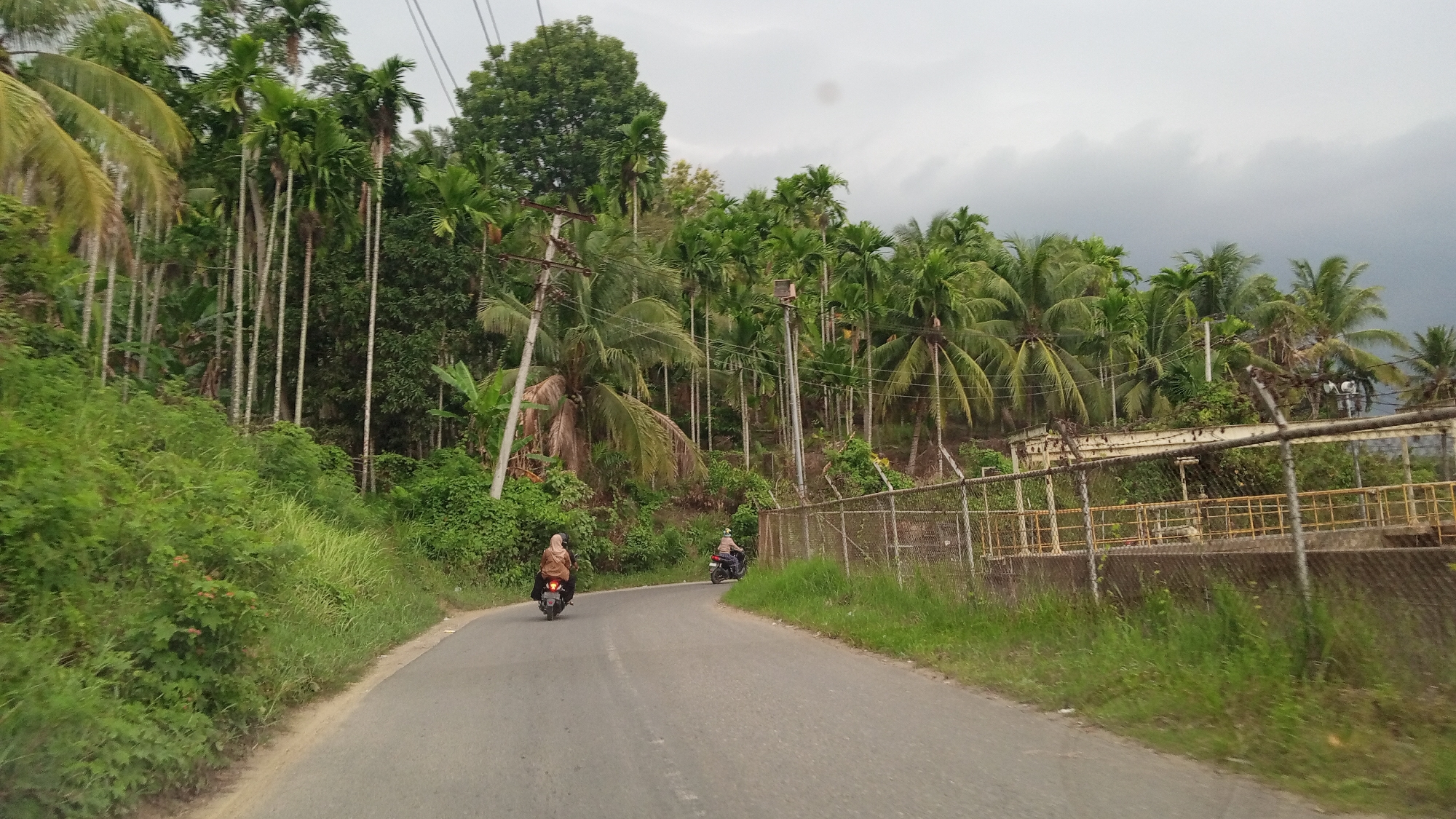 Two individuals on motorcycles are traveling along a paved road that curves through a lush, tropical landscape. Tall, slender palm trees and dense green foliage line both sides of the road. In the background, a chain-link fence encloses a structure with a yellow railing. The sky is overcast, with dark clouds suggesting impending rain. The scene appears to be a rural or semi-rural area, possibly in Blang Mee, Indonesia, given the tropical vegetation and the style of the motorcycles.