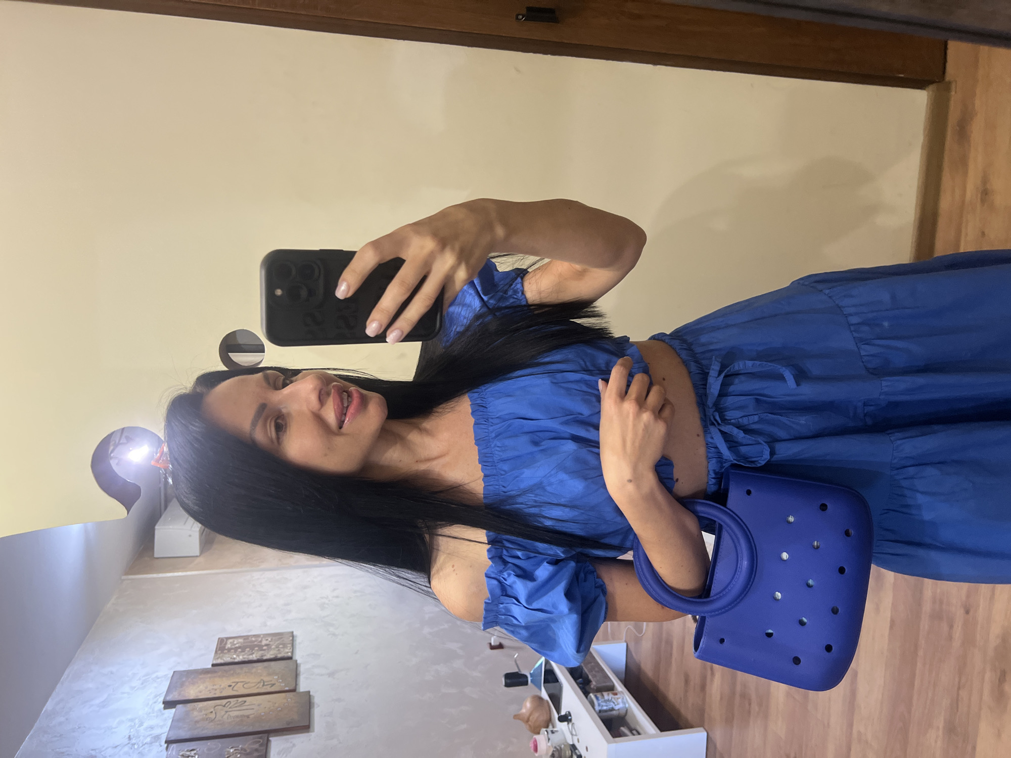 A woman with long, dark hair is standing indoors, taking a mirror selfie. She is wearing a blue off-the-shoulder crop top and a matching tiered skirt. She is holding a blue tote bag with a circular handle and circular perforations. The background includes a wall with decorative panels, a shelf with various items, and a doorway. The lighting suggests an indoor setting, likely a room in a residential property, and the overall aesthetic is casual. The image does not provide any specific visual cues to definitively place the location in Stara Zagora, Bulgaria.
