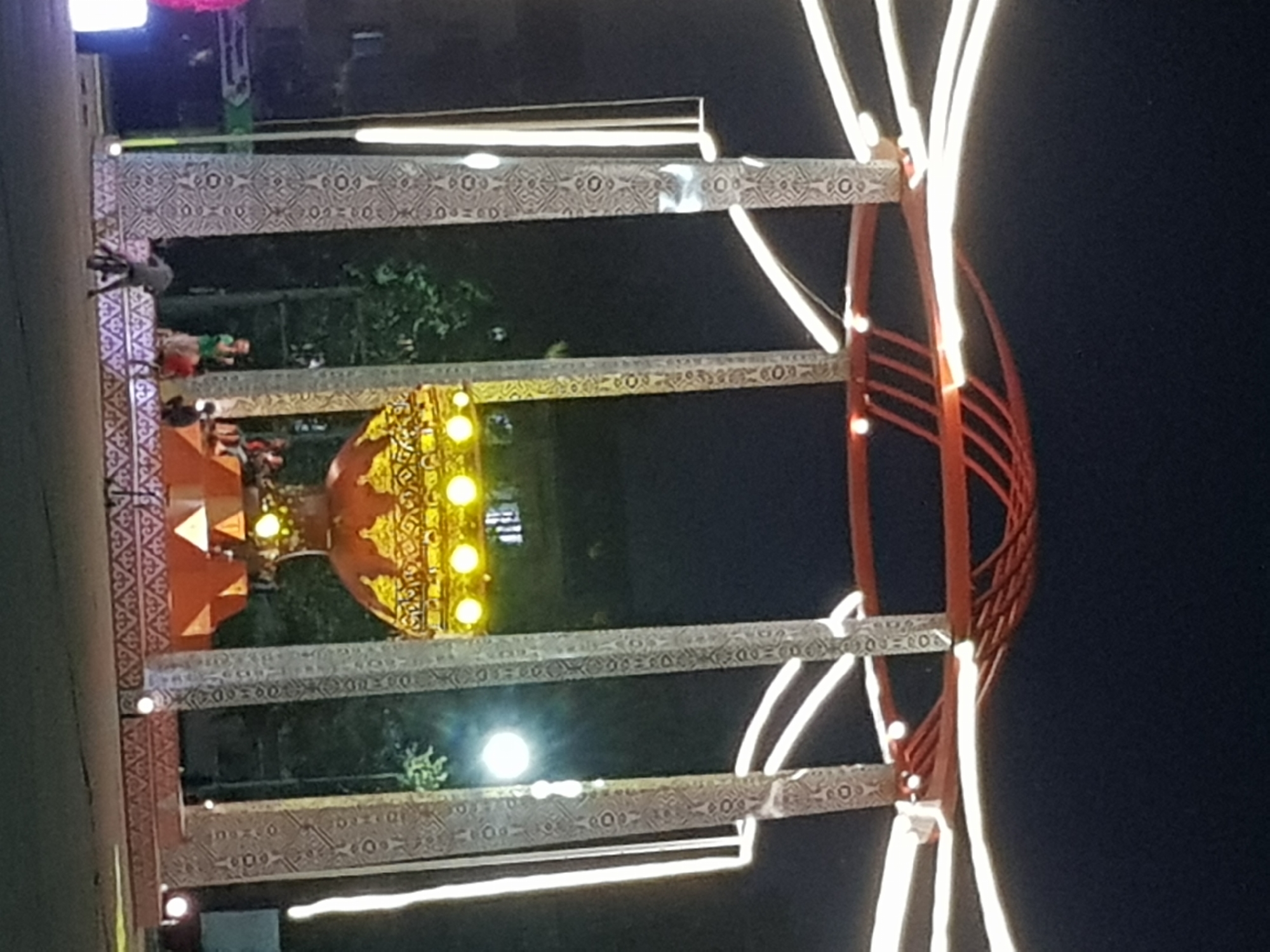 This is a nighttime, outdoor, long-shot photograph of a decorative archway or stage structure. The structure features tall, ornate columns decorated with a repeating geometric pattern. These columns support a circular red canopy. White, curved lights extend outwards from the top of the structure, creating a starburst effect. In the center of the structure, there is a large, ornate, golden bowl-like decoration illuminated by yellow lights. At the base of the structure, a patterned platform is visible, and several people are standing or sitting on it. In the foreground, a person on a bicycle is riding away from the camera. The background shows a dark sky and some trees. The scene appears to be in a public area, possibly a street or plaza. The photograph is blurry and has a grainy quality, which could be due to low light conditions or the quality of the camera used. The exact location in the city of Kurchatov, Kazakhstan, is not discernible from the image itself.