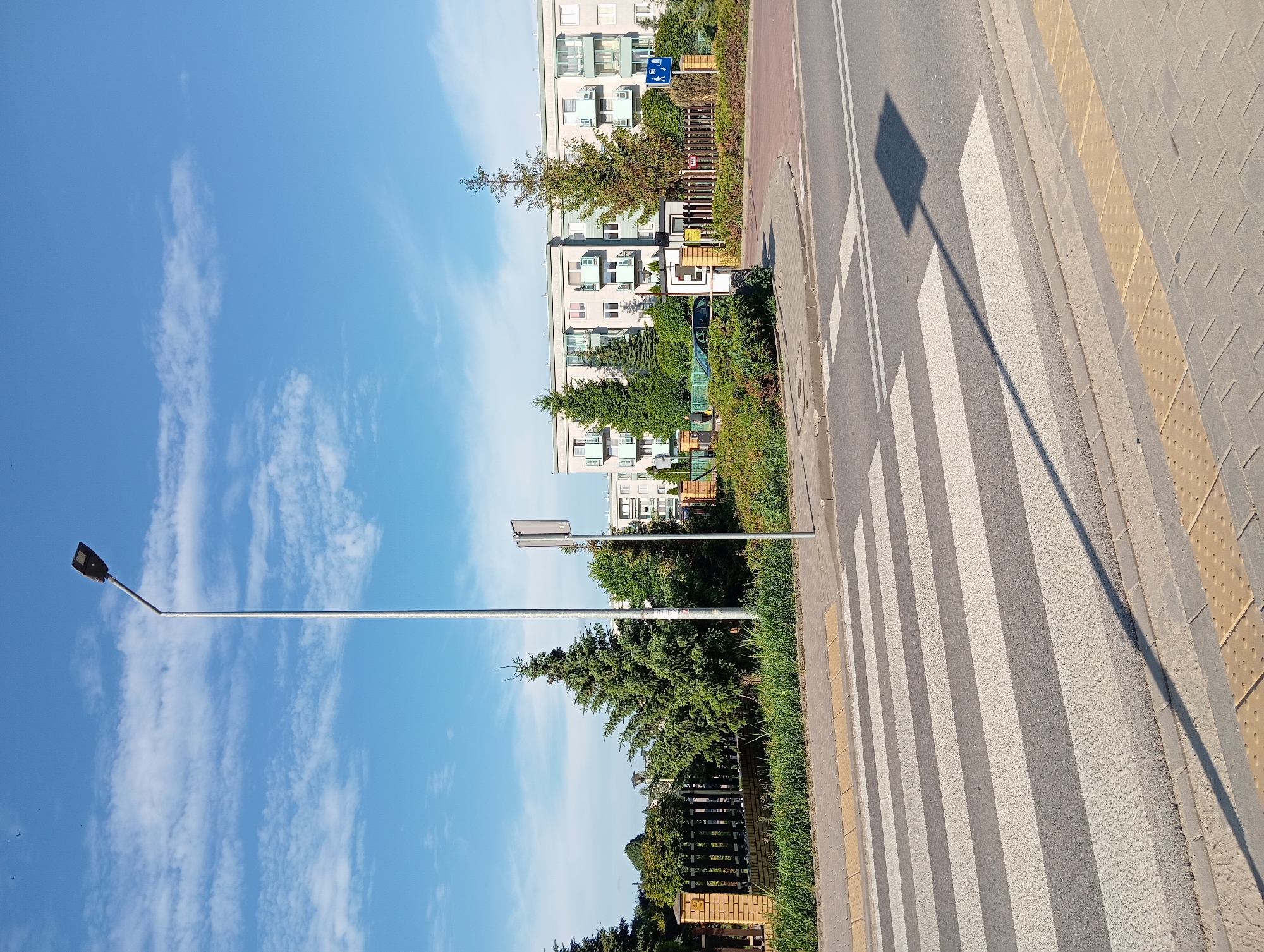 The image shows a street scene with a pedestrian crossing in the foreground. The crossing is marked with white and gray stripes. A street lamp with a dark fixture extends from the left side of the image into the sky, casting a long shadow across the crossing. In the background, there is a multi-story apartment building with balconies. Several evergreen trees are planted in front of the building. A fence, made of brick pillars and dark wooden slats, runs along the edge of the property. The sky is blue with scattered white clouds. The overall environment suggests an urban or suburban setting.