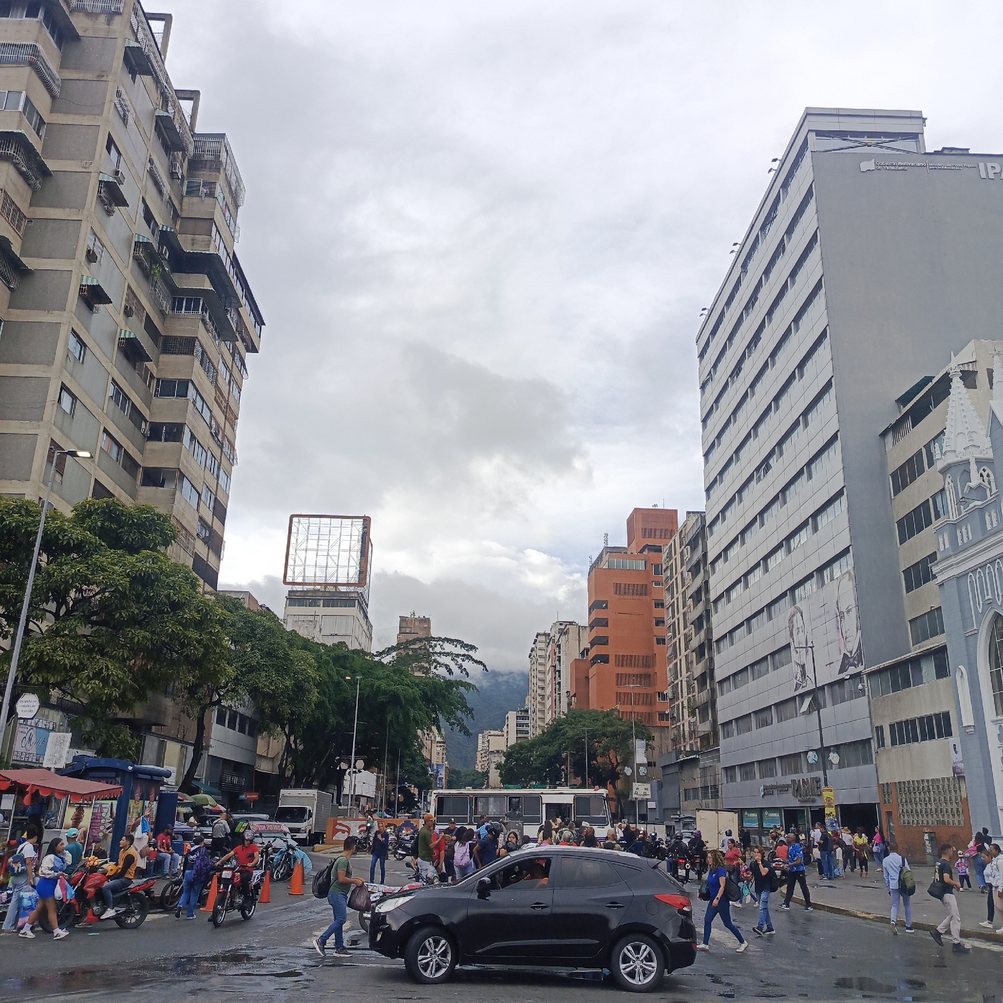This is a wide shot of a street scene in Caracas, Venezuela, under an overcast sky. The image is dominated by tall, multi-story buildings lining both sides of a wide street. On the left, a grey, concrete apartment building with many windows and balconies is prominent, its facade showing signs of age. To the right of this, a large billboard structure stands partially obscured by trees. Further down the street, a tall, modern grey building with many windows displays the text "Gobierno Bolivariano de Venezuela" and "IPA". An ornate, light blue and white building with spires, possibly a church, is visible on the far right.
The street itself is wet, indicating recent rain. A black SUV is crossing the street in the foreground, and several people are walking and standing on the sidewalks and in the street. There are also numerous motorcycles, some parked and some in use. Stalls or small shops are set up along the side of the street, with vendors and customers present. People are dressed in casual clothing, and some are carrying bags. The overall impression is of a busy urban environment.