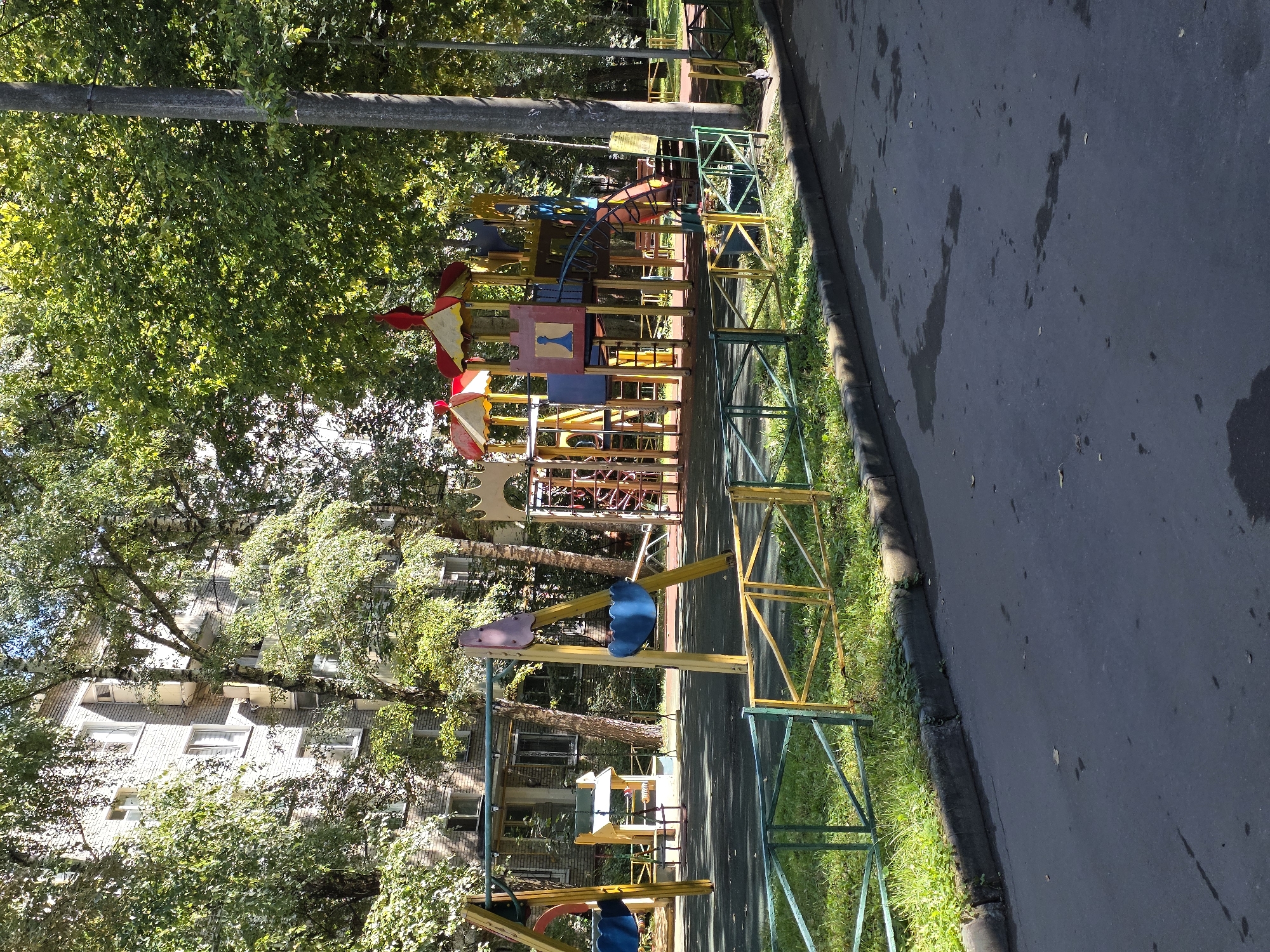 The image shows a children's playground located next to a residential building, likely in an urban environment. The playground features a variety of equipment, including swings with blue seats and a large, multi-colored play structure with slides and climbing elements. The ground surface of the playground is dark and appears to be made of safety matting. A low fence, painted in alternating sections of green and yellow, surrounds the playground. Beyond the fence, there is a strip of grass and then an asphalt road. In the background, a multi-story brick building with multiple windows and balconies is visible, partially obscured by lush green trees. The overall scene suggests a typical neighborhood playground, likely in Moscow, Russia, given the architectural style of the building and the presence of trees.