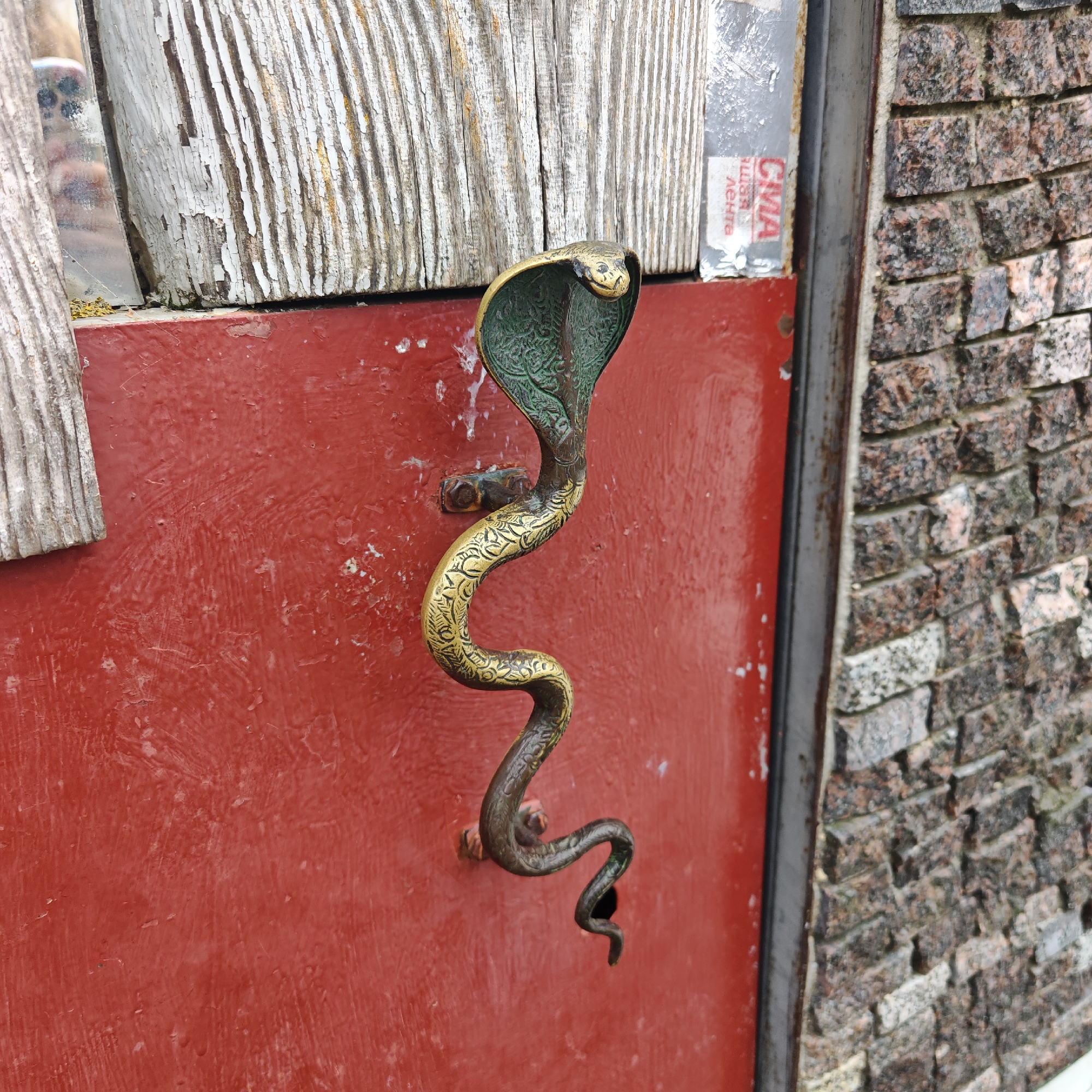 A medium, eye-level shot shows a cobra-shaped door handle on a red door with peeling paint. The handle is made of brass, with detailed engravings along the snake’s body. The cobra’s head is raised and flared, with a green interior. The handle is attached to the door with visible screws.
To the left of the door, there is a weathered wooden surface with peeling white paint. To the right, there is a stone wall made of small, dark-colored bricks. At the top of the door is a sticker with the letters "CMA" and other Cyrillic text.
Based on the availability of this style of architecture and door handles, it is suspected that this photo was taken in Rostov, Russia.
