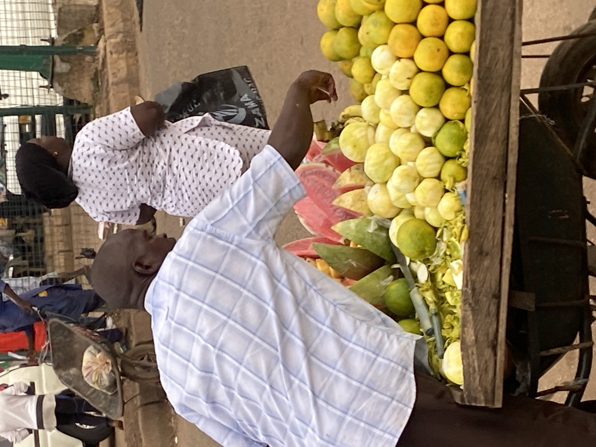 The image captures a street scene in Bukuru, Nigeria, with a focus on a fruit vendor and his cart. A man wearing a blue and white checkered shirt is bending over, reaching towards a cart laden with various fruits, including what appear to be watermelons, limes, and lemons. The cart is rustic, constructed from wood and metal, and is part of a larger wheeled mechanism. Behind the vendor, a woman in a white patterned outfit and a black headscarf is visible, holding a dark bag. In the background, other people and a white vehicle are partially discernible, suggesting a bustling urban environment. The ground is dusty, indicative of a dry, possibly hot climate. The scene depicts a typical street market scenario, with the vendor actively engaged in his trade.