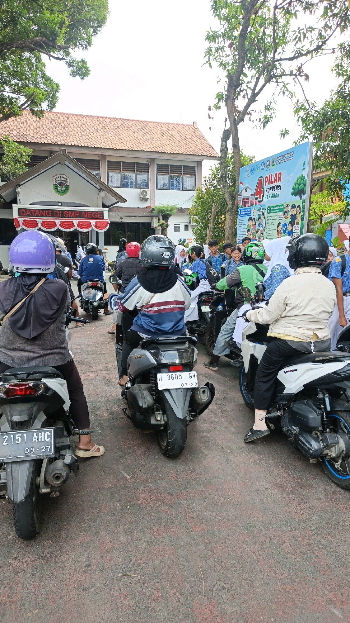 The image depicts a scene outside a school in Sidomulyo, Indonesia. In the foreground, several people are on motorcycles, with many appearing to be students or staff. The building in the background is a school with red-tiled roofs and visible windows. A banner with the text "4 PILAR KONVENSI HAK ANAK" (4 Pillars of the Convention on the Rights of the Child) is displayed on the right side of the image. The ground is paved and appears to be a schoolyard or parking area. The atmosphere suggests a typical school day, possibly during arrival or dismissal, with many people using motorcycles for transportation.