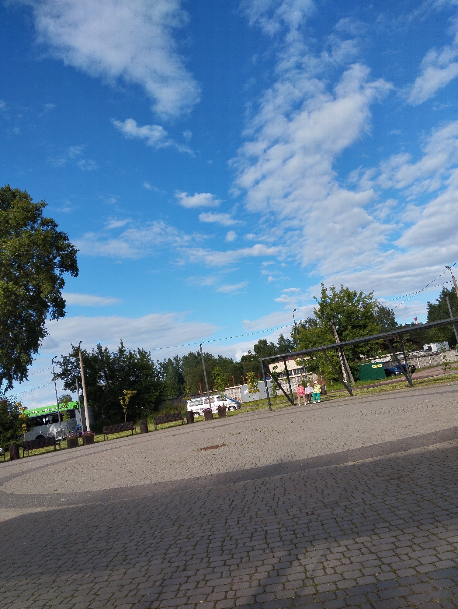 The image shows a paved area with a circular pattern in the foreground. In the middle ground, there is a black metal canopy structure with three people sitting beneath it, one in a pink jacket, one in a yellow jacket, and one in a green jacket. To the left of the canopy, there is a white bus with green lettering on its side, parked near a bench. Beyond the bus and canopy, there are trees and a building in the background. The sky is blue with white clouds. The image is taken at street level and appears to be a public space in the city of Kingisepp, Russia.