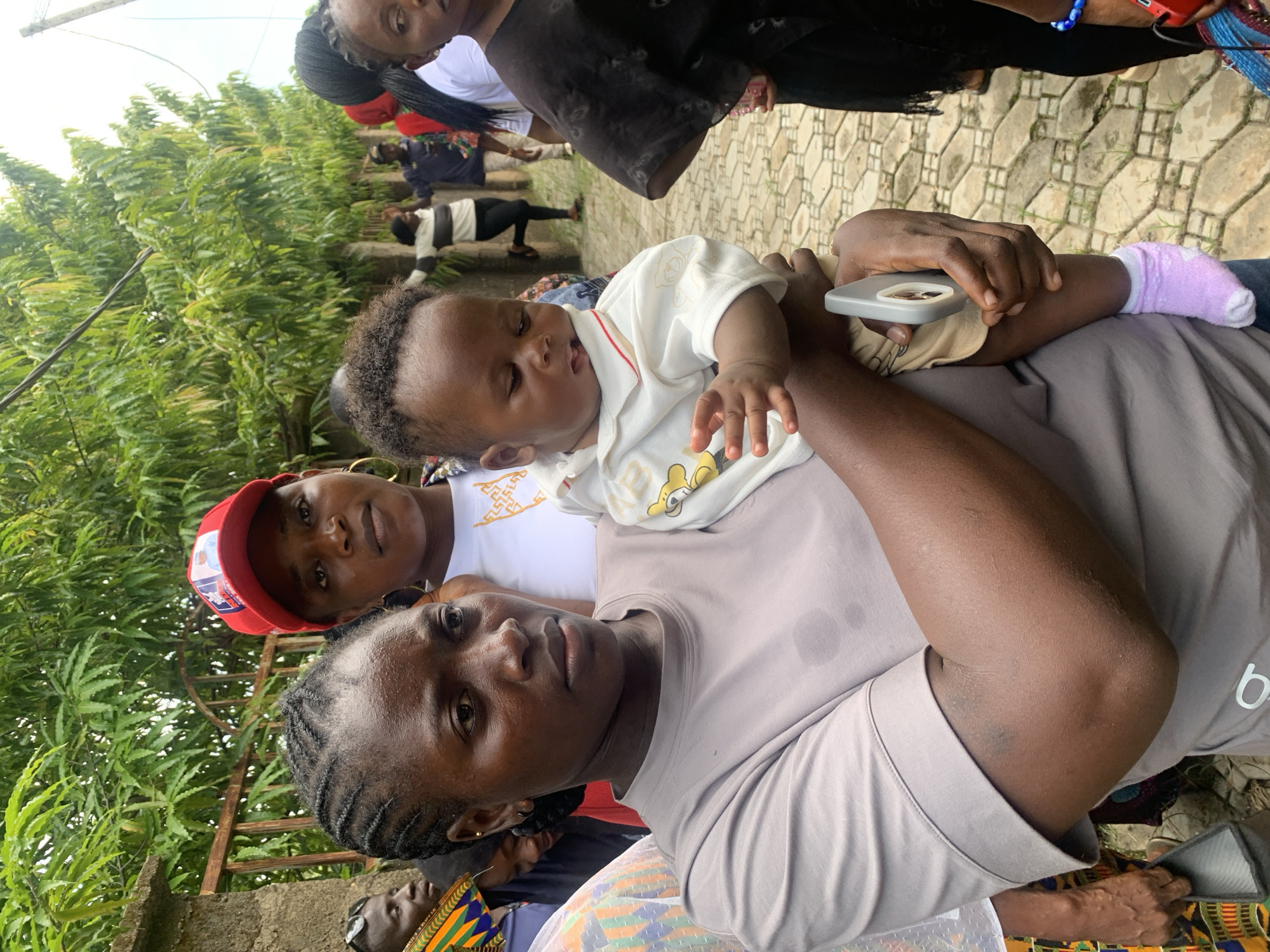In Akwanga, Nigeria, an outdoor scene features a group of people standing on a paved walkway bordered by a metal fence and lush green trees. In the foreground, a woman with dark skin and braided hair holds a baby. She wears a grey t-shirt. The baby, dressed in a white shirt with a teddy bear design, is held in her arms, with one hand resting on the baby's back. A person wearing a red cap stands behind and to the left of the woman. Other individuals are visible in the background, some walking and others standing. The environment suggests a public or residential outdoor area.