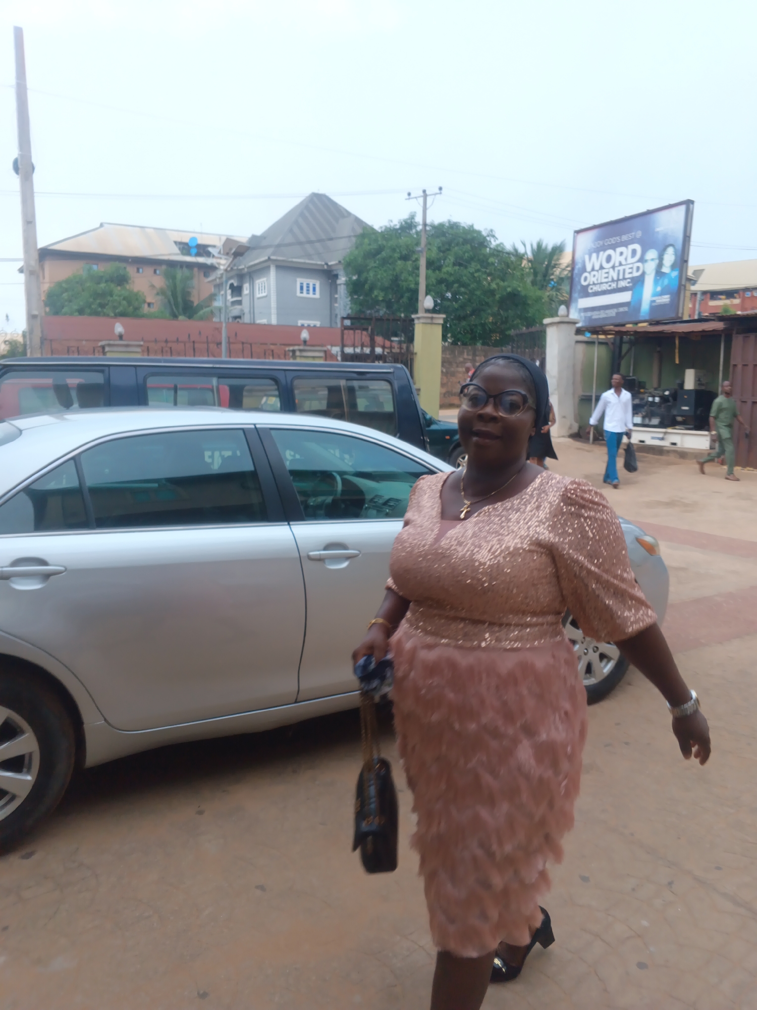 In Nkpor, Nigeria, a woman in a rose gold sequined top and feathered skirt walks beside a silver sedan. She wears glasses and a black headscarf, carrying a dark handbag. In the background, several buildings, including one with a billboard advertising "WORD ORIENTED CHURCH INC.", and two men, one in a white shirt and blue pants, and another in a green shirt and pants, are visible. The ground is paved with reddish-brown earth.