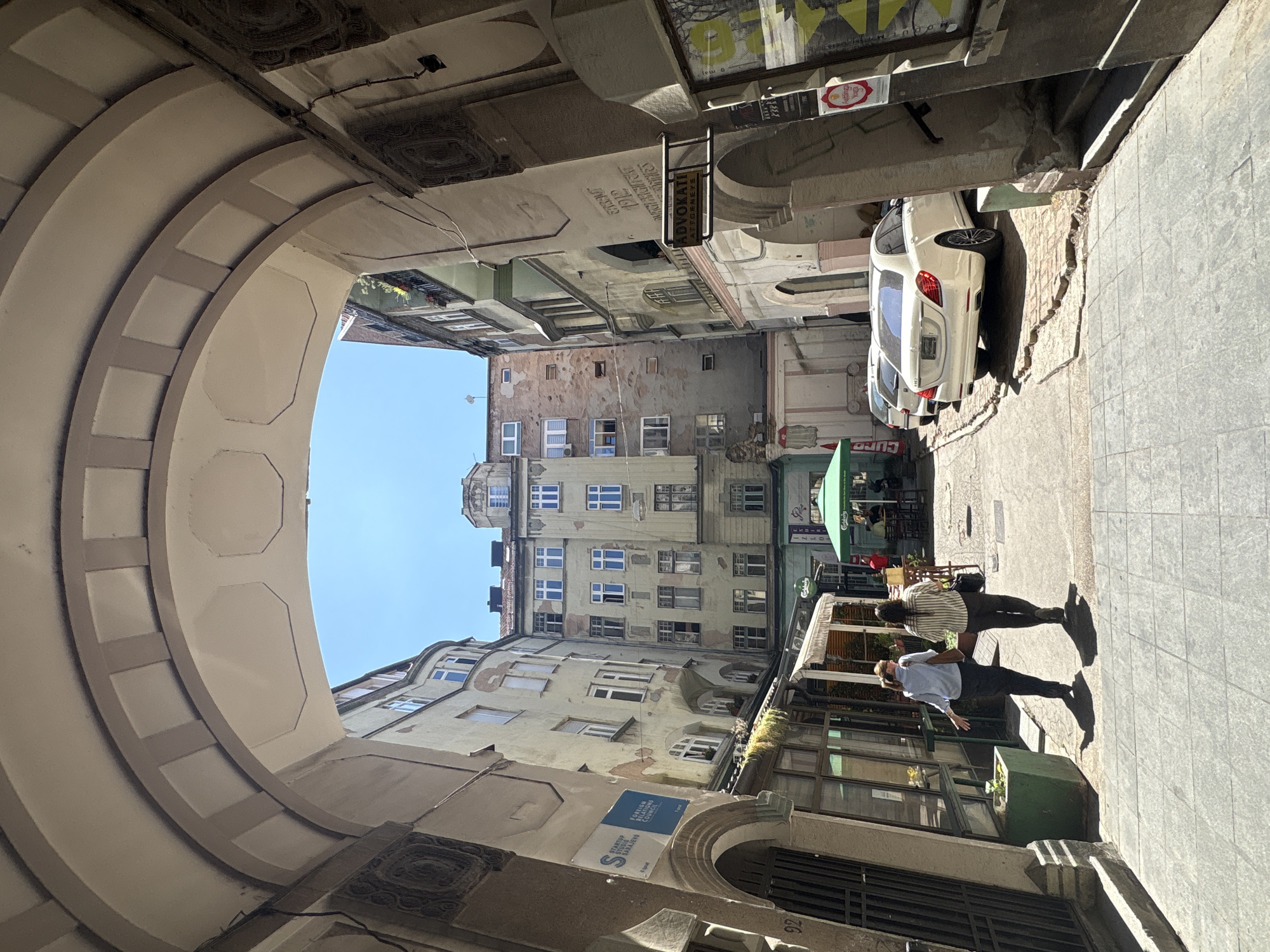 This image captures a sunlit, narrow European-style alleyway in Sarajevo, Bosnia and Herzegovina. The perspective is from beneath a large, ornate archway, looking towards a cluster of buildings. The buildings are a mix of architectural styles, with aged facades, numerous windows, and balconies.
In the foreground, two women are walking away from the camera down the cobblestone alley. The woman on the left wears a light blue shirt and dark pants, carrying a brown bag. The woman on the right is dressed in a striped shirt and dark pants, with a black bag over her shoulder. Their shadows are cast long on the ground.
Further down the alley, several cars are parked. A white Mercedes sedan is prominently visible, facing away from the viewer. Another white car is parked in front of it. A green umbrella shades an outdoor seating area, suggesting a cafe or restaurant. Various signs are visible on the buildings, including one that reads "ADOKATI ATTORNEYS." A blue sign on a building to the left indicates "STARTUP STUDIO SARAJEVO" and "FOREIGN RELATIONS COUNCIL." The sky is clear and blue. The overall impression is one of a historic urban environment on a bright day.