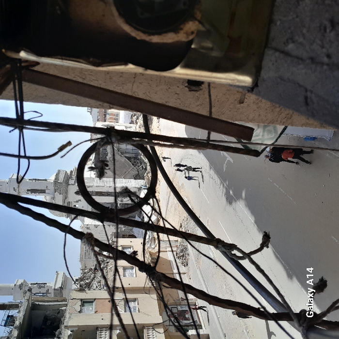 An elevated view captures a street scene in the city of Khan Yunis, Palestinian Territory, characterized by extensive structural damage under a clear daytime sky. In the immediate foreground, a dense tangle of dark, rusted metal rebar and wires, likely from a destroyed structure, frames the composition. To the far right, a concrete element and a metallic object are partially visible.
The street below is a paved roadway, appearing somewhat uneven with debris. Along the left side of the street, multiple multi-story residential or commercial buildings exhibit severe damage, with large sections of their facades and interiors collapsed, exposing debris and rubble. In the background, a prominent mosque-like structure with a white minaret stands partially destroyed, showing significant breaches and missing sections in its upper levels and dome. Other damaged buildings are visible behind it.
On the street, several individuals are present. In the mid-ground, a group of approximately five people walks along the paved road. Two individuals at the center of this group are observed jointly carrying a long, light-colored object, possibly a pipe or beam. Further down the street on the left sidewalk, a lone individual in a dark shirt walks away from the camera. Closer to the right foreground, another small group of three to four individuals is seen standing on the street or sidewalk; one person is wearing an orange shirt, while others wear dark clothing. All individuals appear to be civilians.